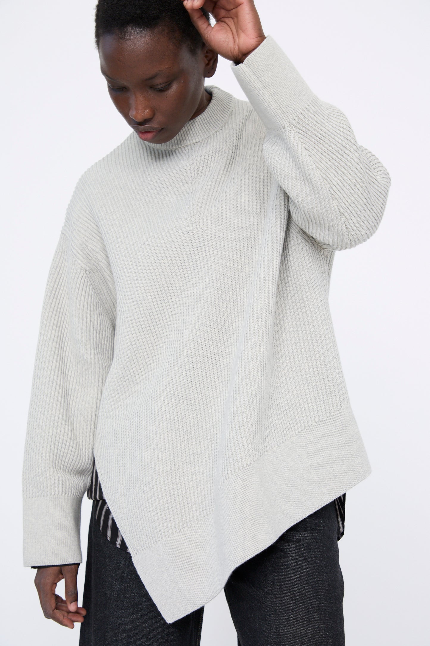 A person is wearing the Studio Nicholson Dry Cotton Rib Verda Asymmetric Knit in Grey Marl and dark pants, standing against a plain white background, with one arm raised. 