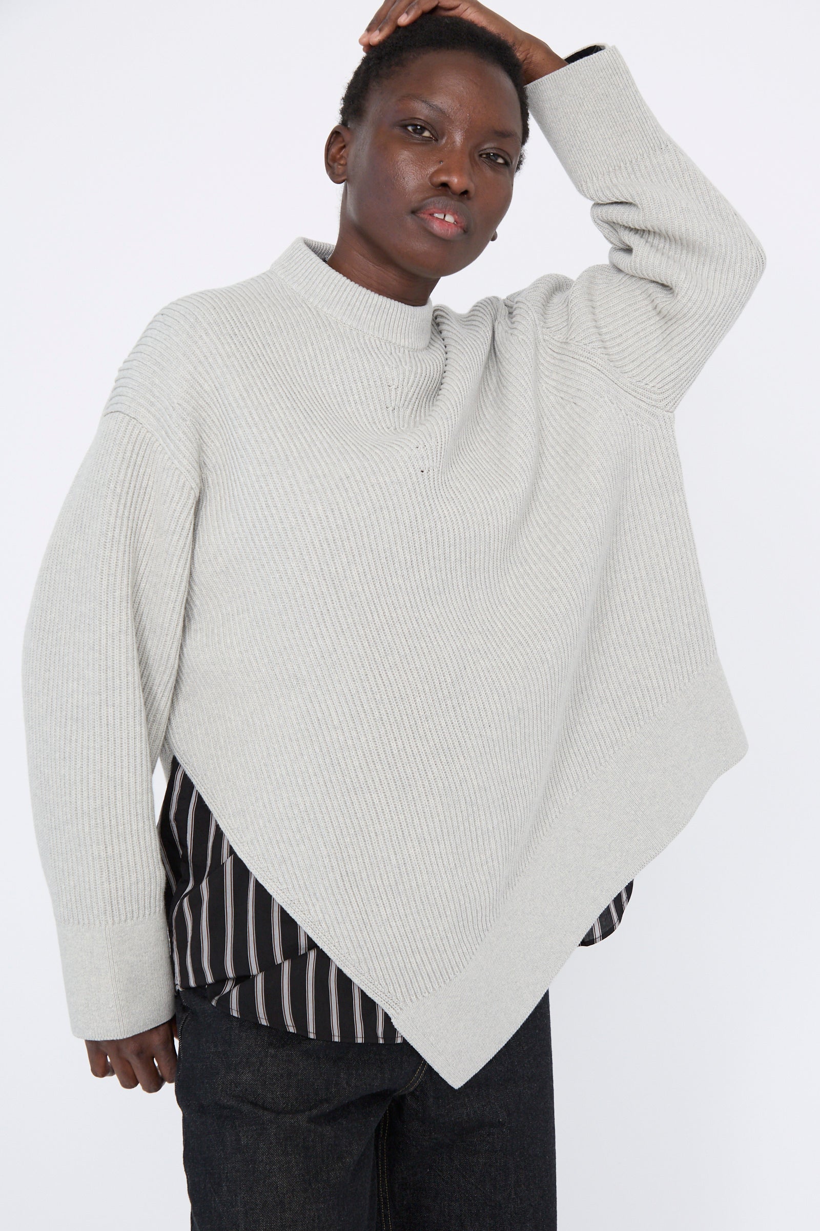 A person poses with one hand on their head, wearing Studio Nicholson's Dry Cotton Rib Verda Asymmetric Knit in Grey Marl over a striped shirt against a plain background.