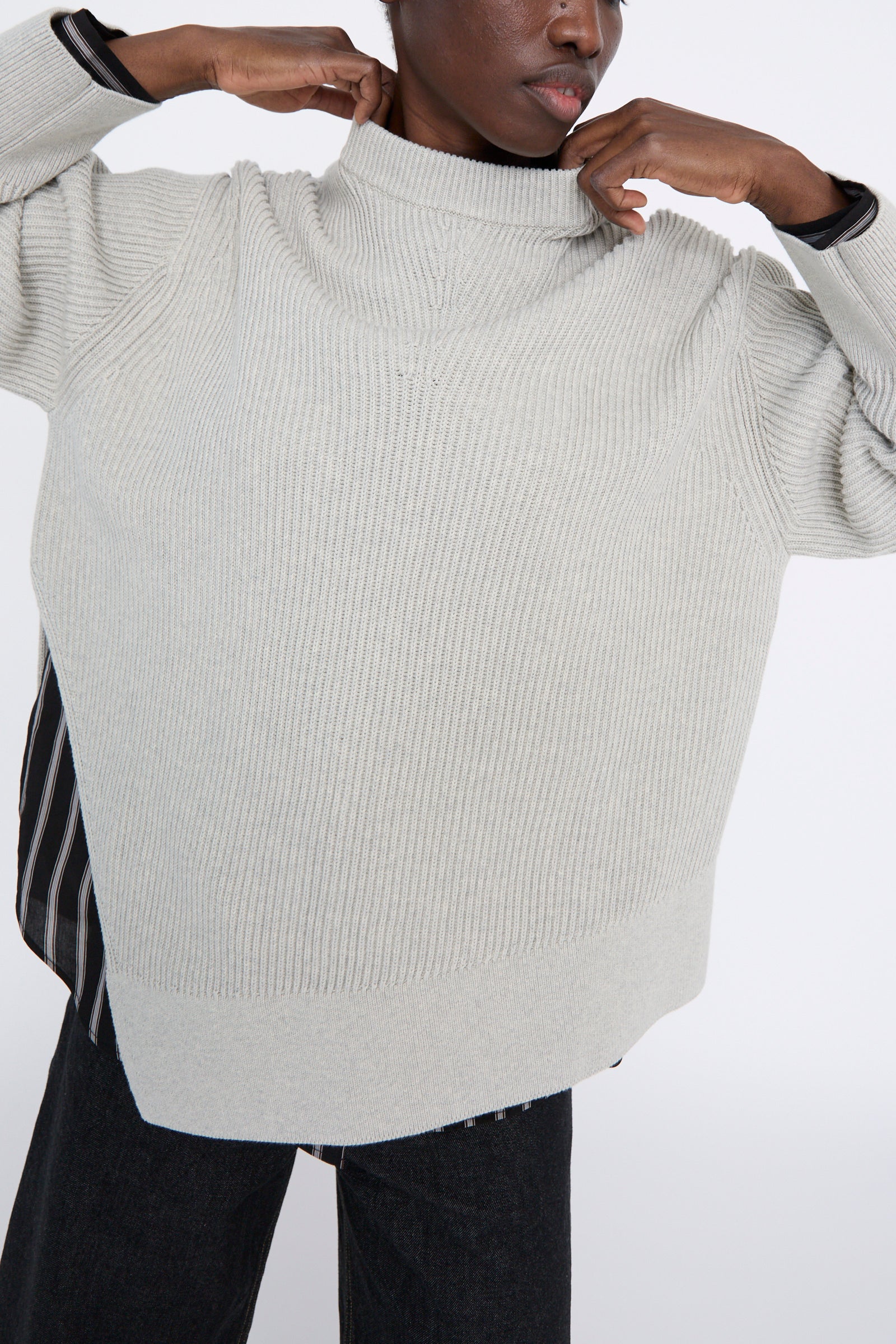 A person adjusts the oversized Studio Nicholson Dry Cotton Rib Verda Asymmetric Knit in Grey Marl, layered over a striped shirt against a neutral background.