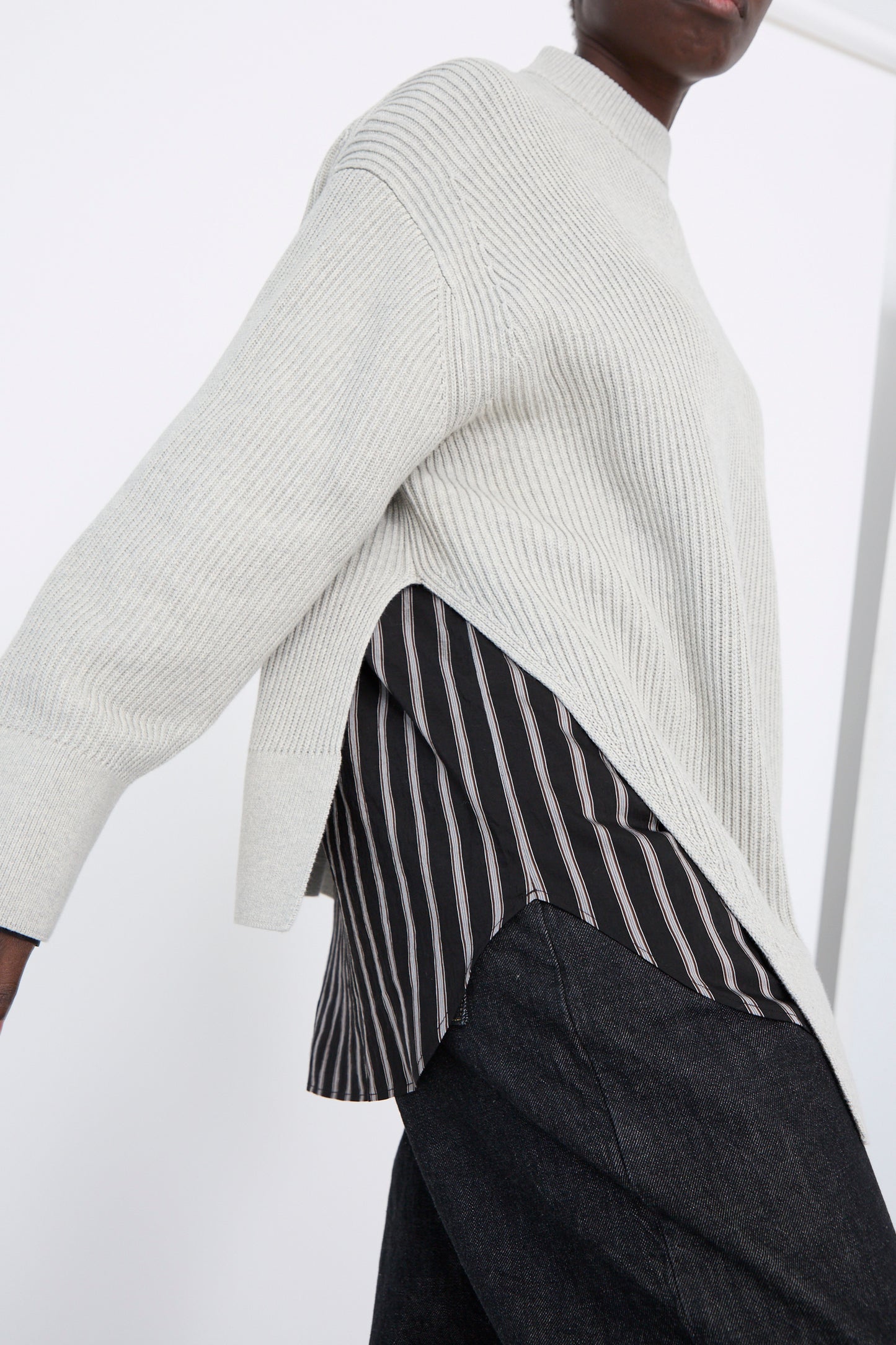 A person wears the oversized Dry Cotton Rib Verda Asymmetric Knit in Grey Marl by Studio Nicholson over a black and white striped shirt, paired with dark pants.