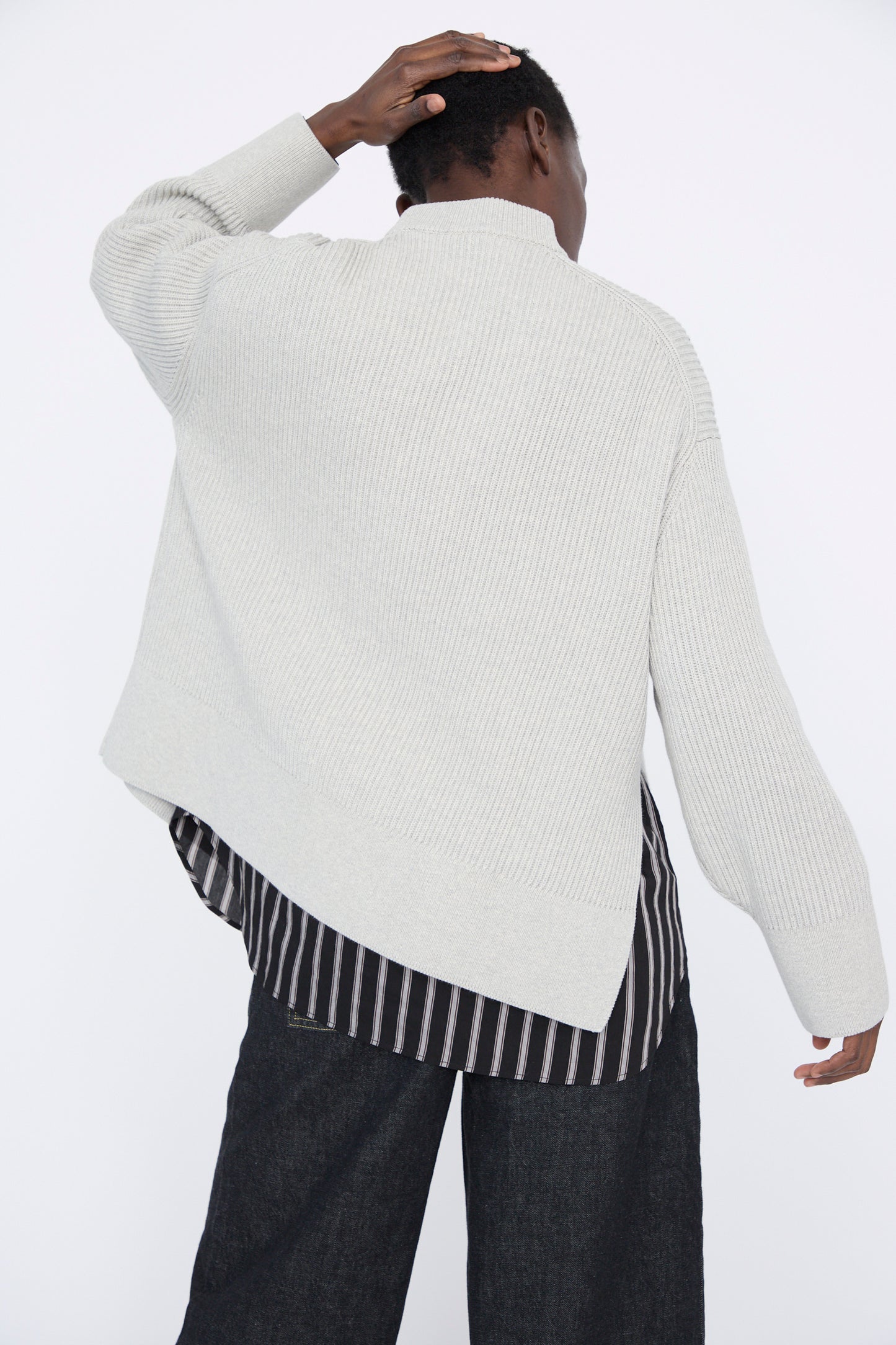 Wearing Studio Nicholson's Dry Cotton Rib Verda Asymmetric Knit in Grey Marl over a striped shirt and dark pants, a person stands against a stark white background with their back turned and hand touching their head.