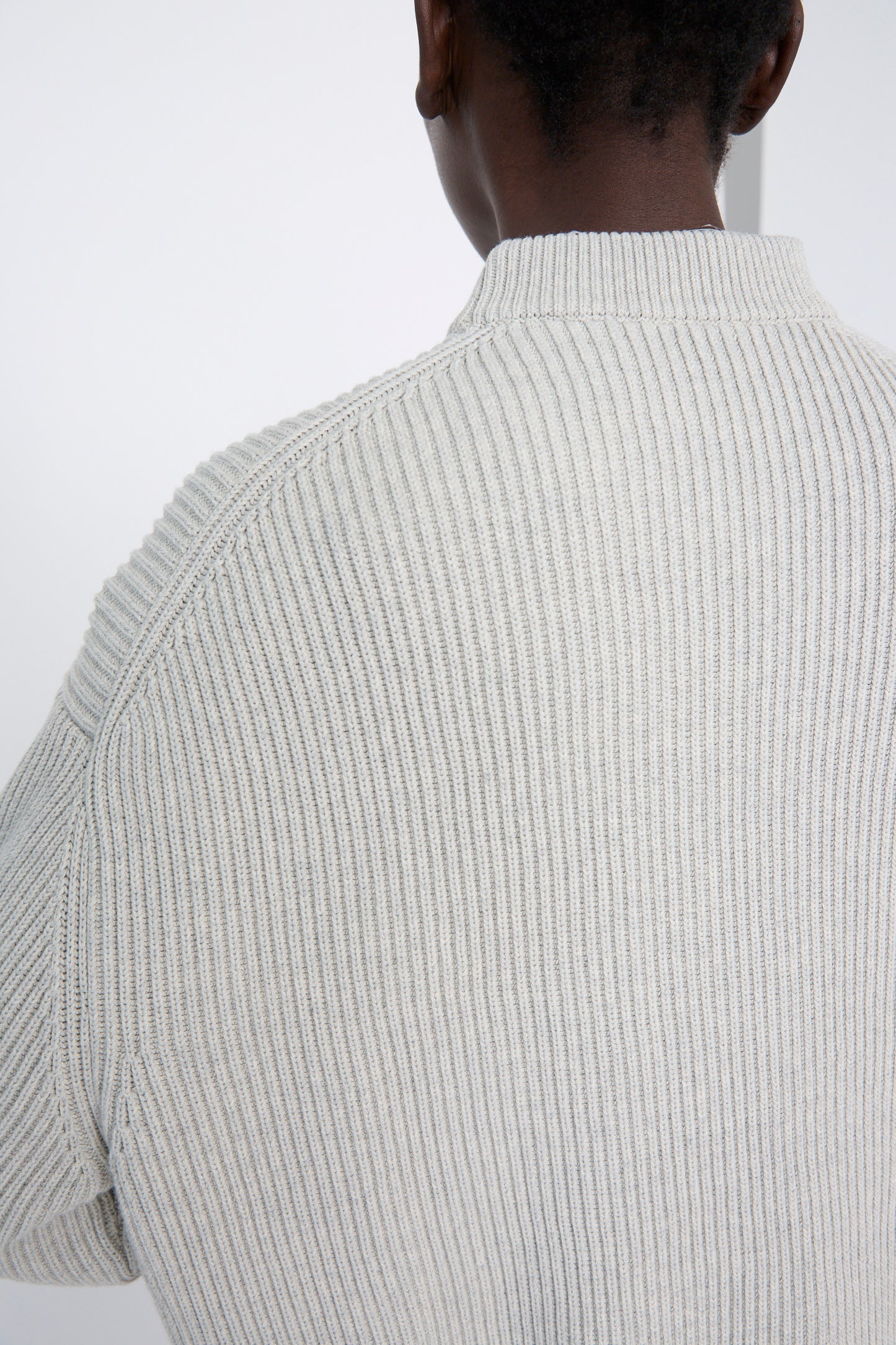 A person wearing Studio Nicholson's Dry Cotton Rib Verda Asymmetric Knit in Grey Marl, viewed from the back, stands against a white background.