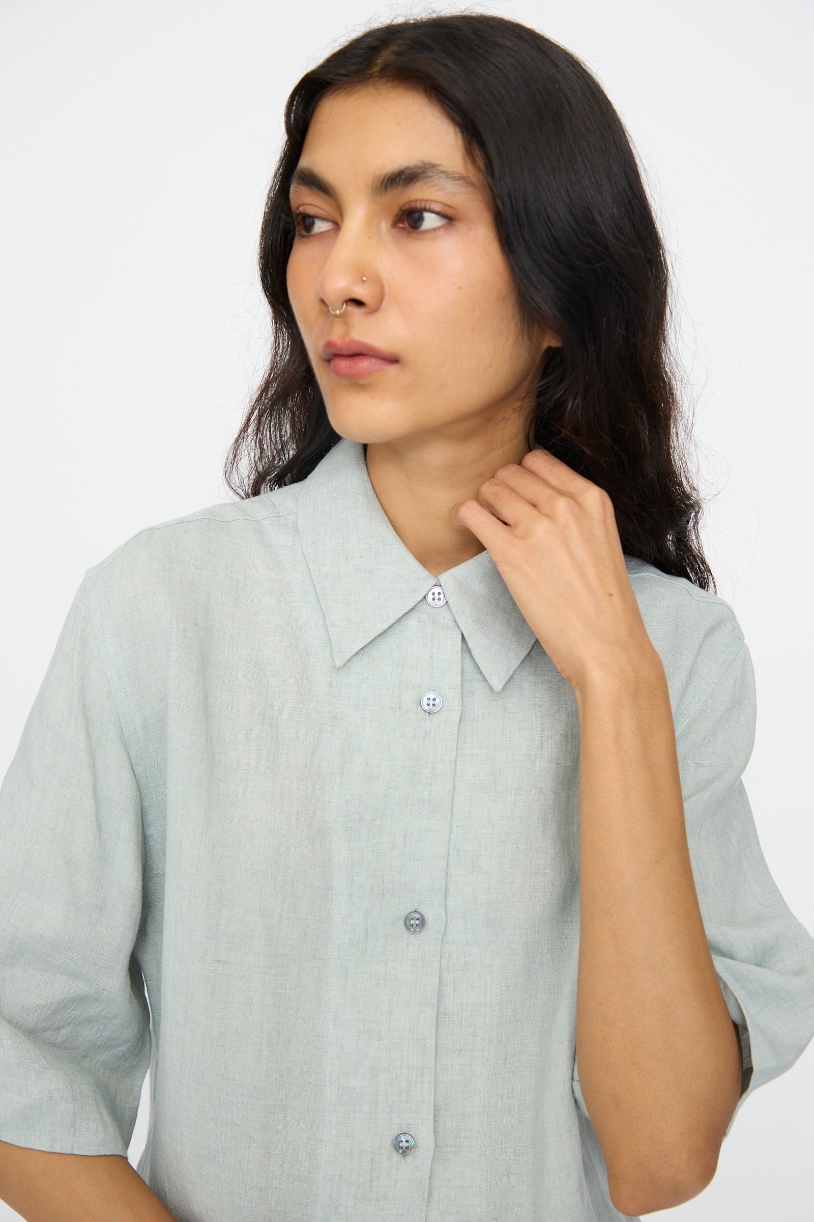 A person with long dark hair wears the Studio Nicholson Estes Shirt in Spearmint, a light gray, short sleeve linen shirt with a relaxed fit, looking to the side and touching their collar.