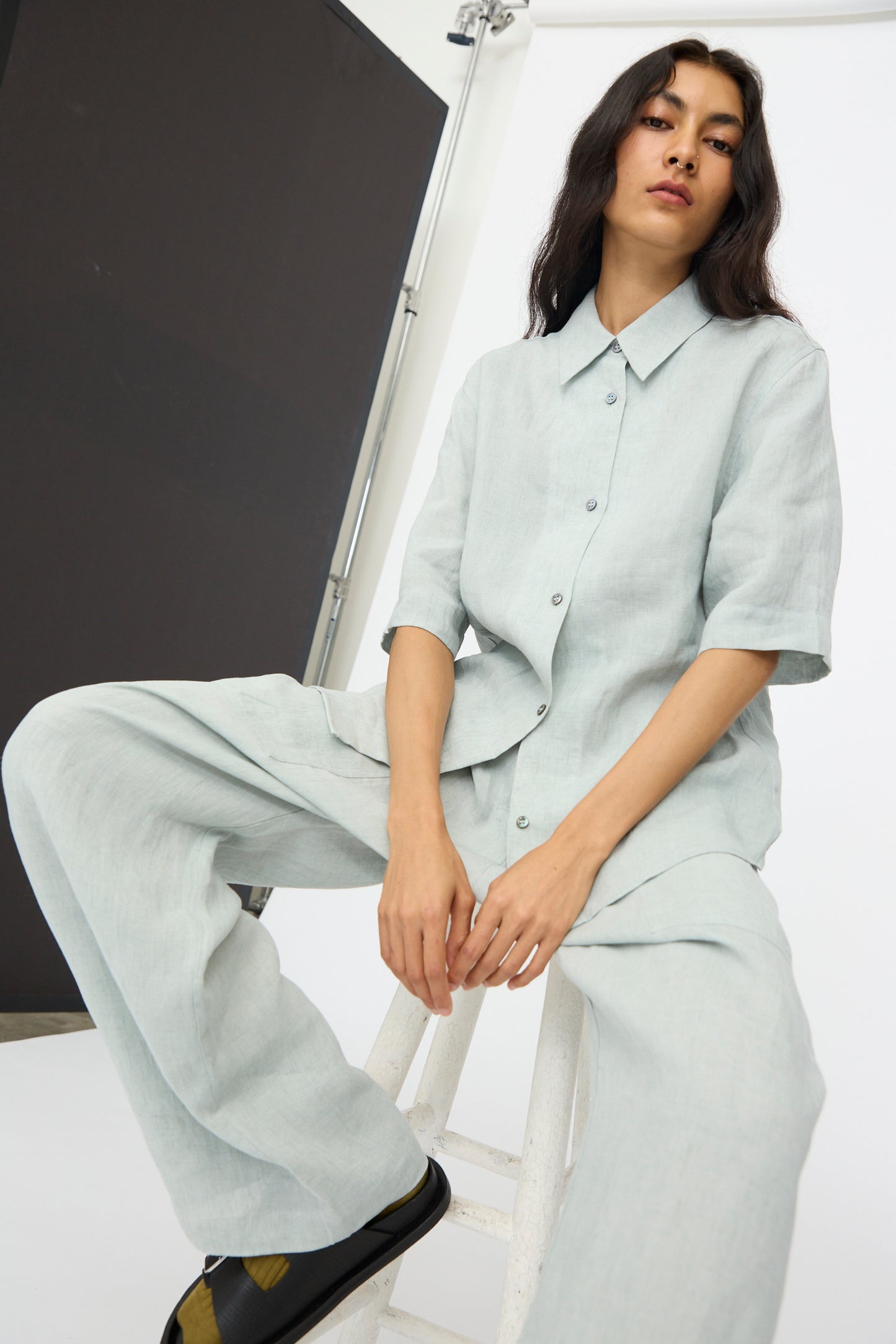 A person sits on a white stool in a studio with black and white backgrounds, wearing Studio Nicholson’s Estes Shirt in Spearmint with matching wide-leg pants and black shoes.