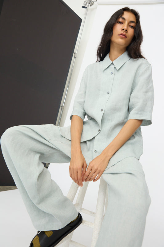 A person sits on a white stool in a studio with black and white backgrounds, wearing Studio Nicholson’s Estes Shirt in Spearmint with matching wide-leg pants and black shoes.