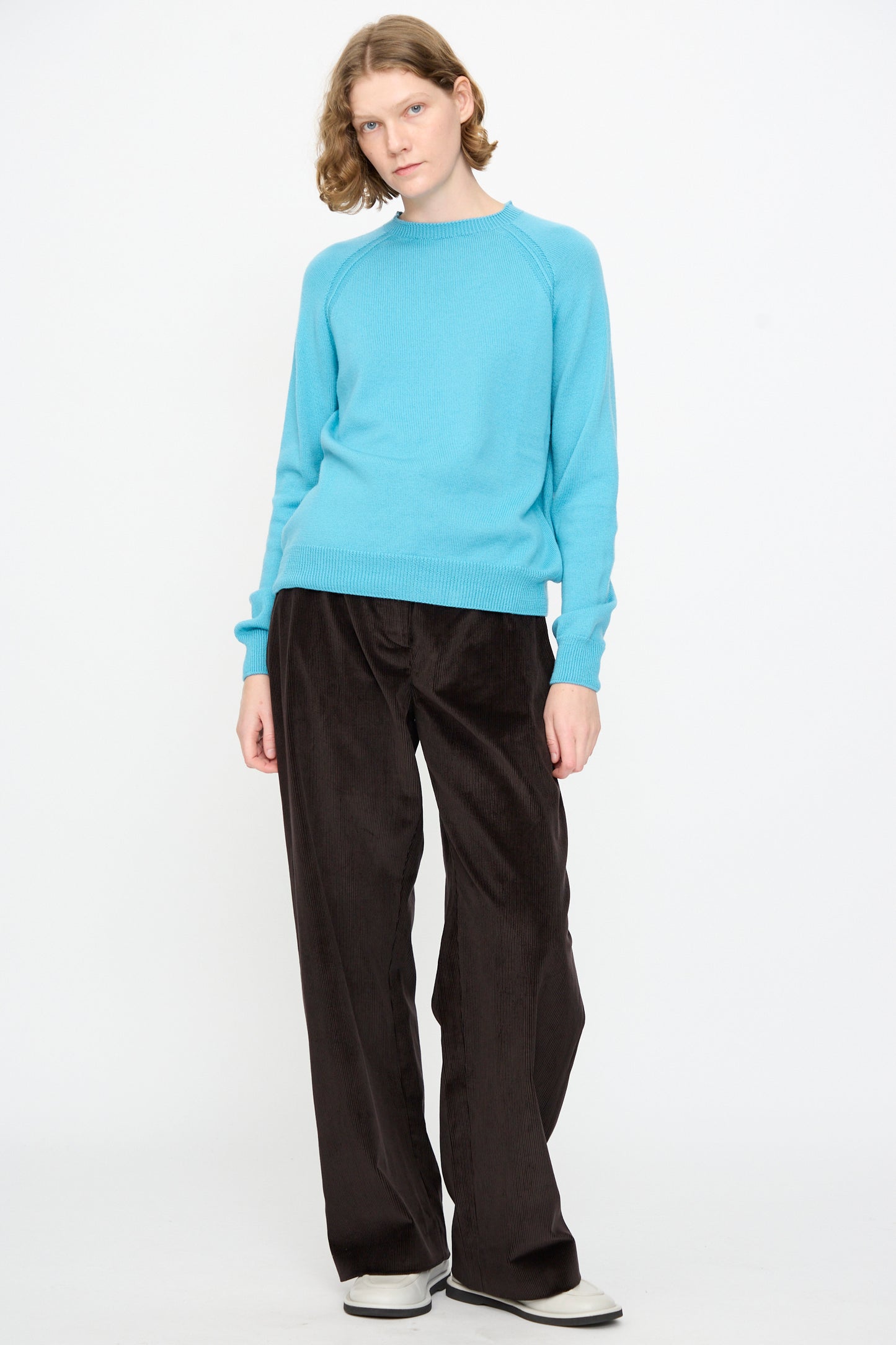 A person wears the Studio Nicholson Extrafine Merino Leonne Sweater in Sky, styled with black wide-leg pants and white shoes, standing against a plain white background.