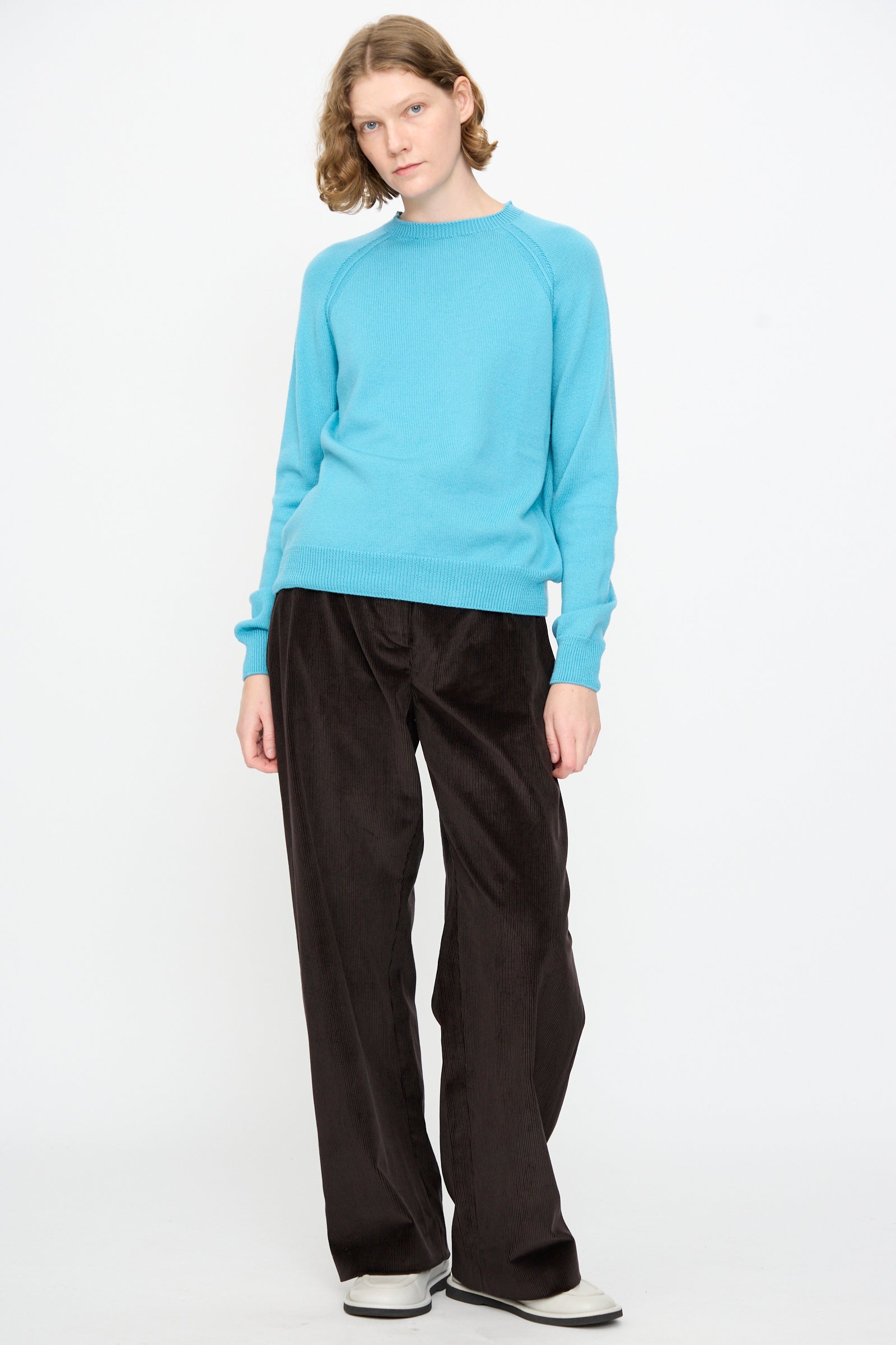 A person wears the Studio Nicholson Extrafine Merino Leonne Sweater in Sky, styled with black wide-leg pants and white shoes, standing against a plain white background.