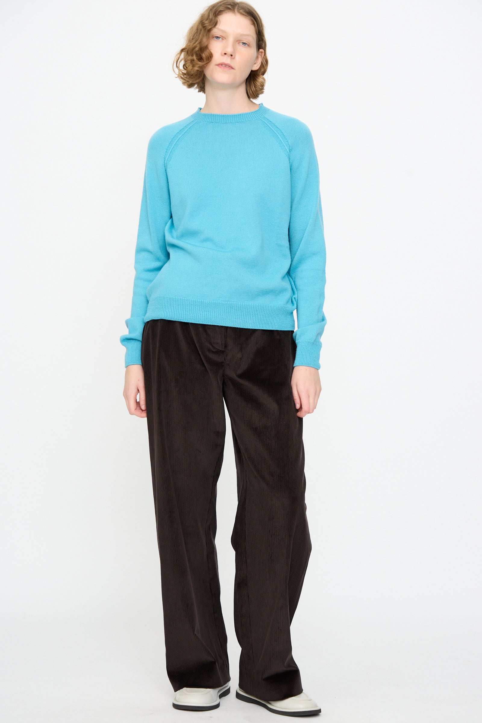 A person stands against a white background, dressed in the Studio Nicholson Extrafine Merino Leonne Sweater in Sky, paired with loose dark brown pants and white sneakers.