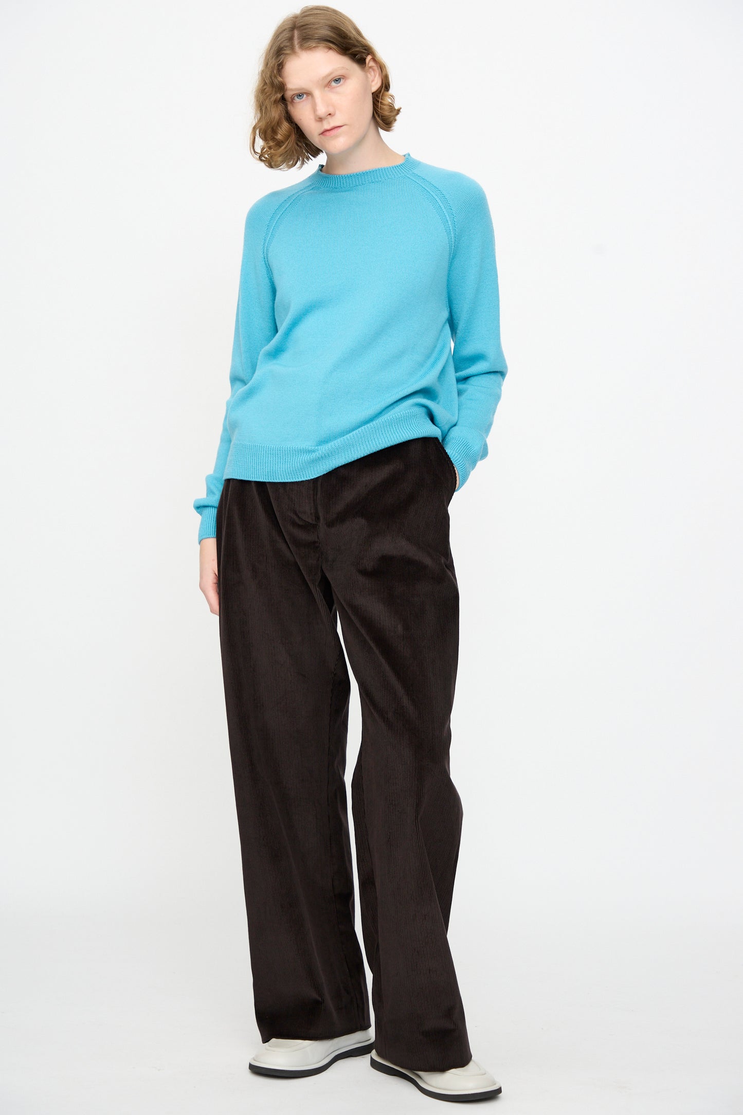 A person stands against a white background, wearing the Studio Nicholson Extrafine Merino Leonne Sweater in Sky—a light blue, 1960s-inspired jumper—paired with dark wide-leg pants and white shoes, with one hand in their pocket.