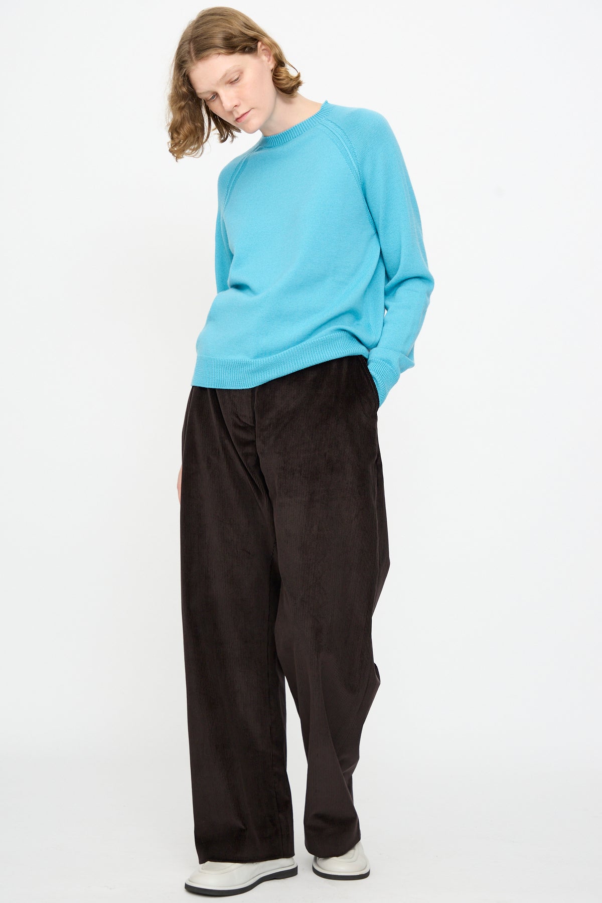 A person stands against a white background, wearing the Studio Nicholson Extrafine Merino Leonne Sweater in Sky, styled with loose black pants and white shoes, hands in pockets and head slightly tilted down.