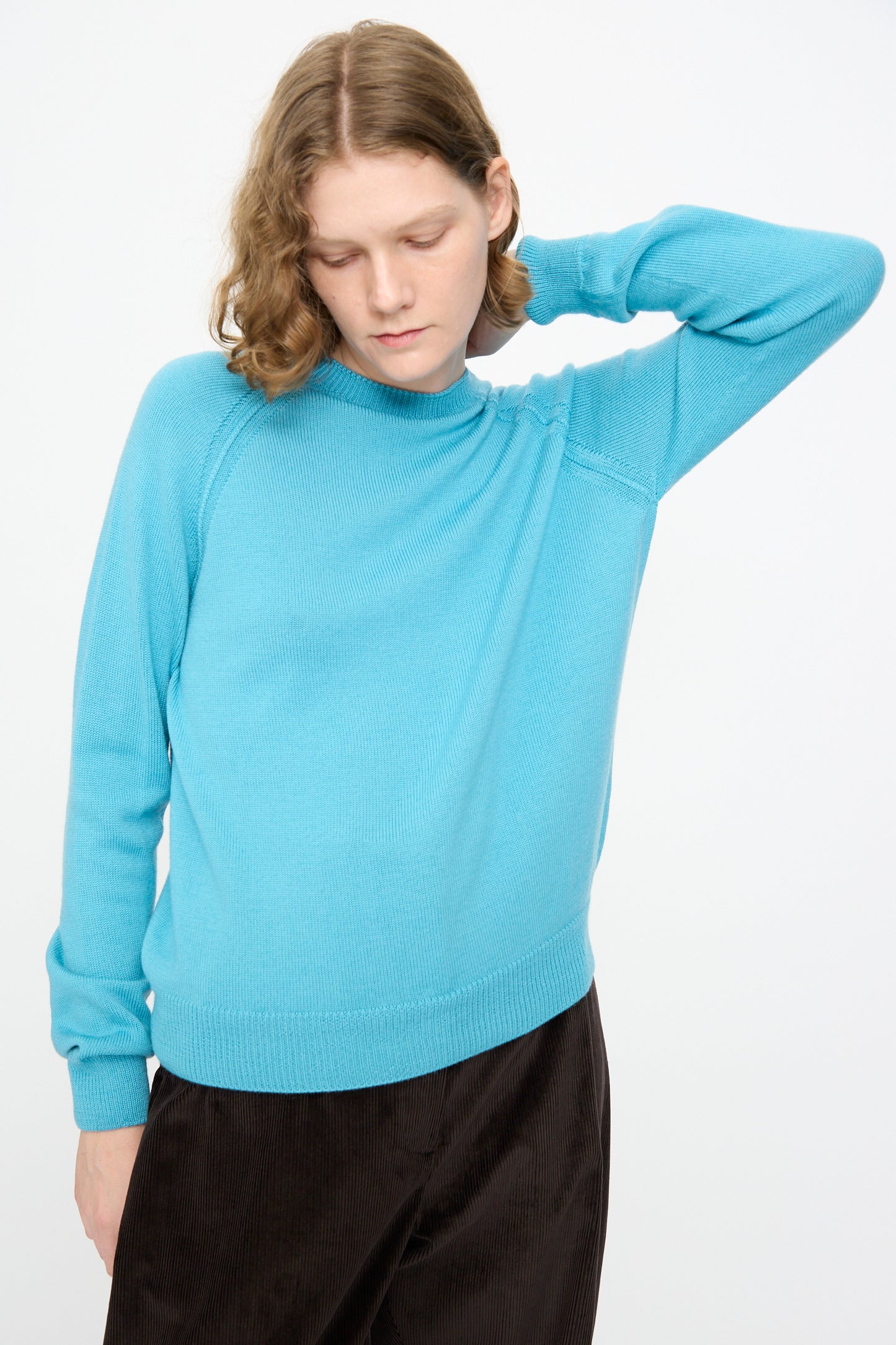 A person wearing the Studio Nicholson Extrafine Merino Leonne Sweater in Sky and dark pants stands against a plain white background, looking down with one hand behind their neck. This 1960s-inspired jumper is made in the UK.