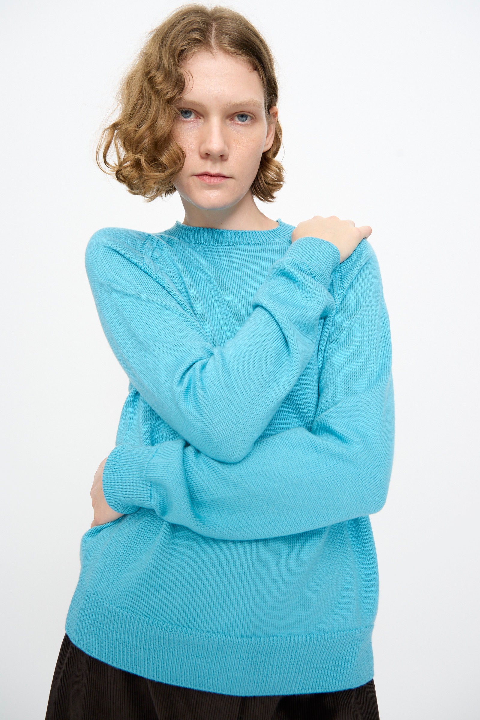 A person with short, curly hair stands against a plain white background, crossing one arm over their chest while wearing the Studio Nicholson Extrafine Merino Leonne Sweater in Sky.