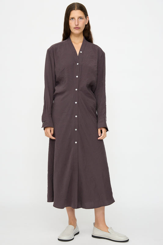 A woman faces forward wearing the Studio Nicholson Fiore Shirt Dress in Yam—an ankle-length, long-sleeve, button-down style—paired with white slip-on shoes against a plain white background.