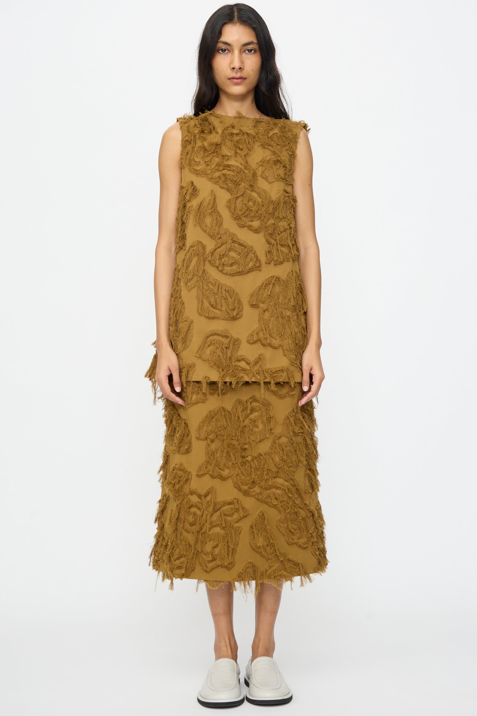 A woman wears the Studio Nicholson Fringe Jacquard Mager Top in Mustard with a matching midi skirt featuring textured fringe, styled with white slip-on shoes against a plain white background.