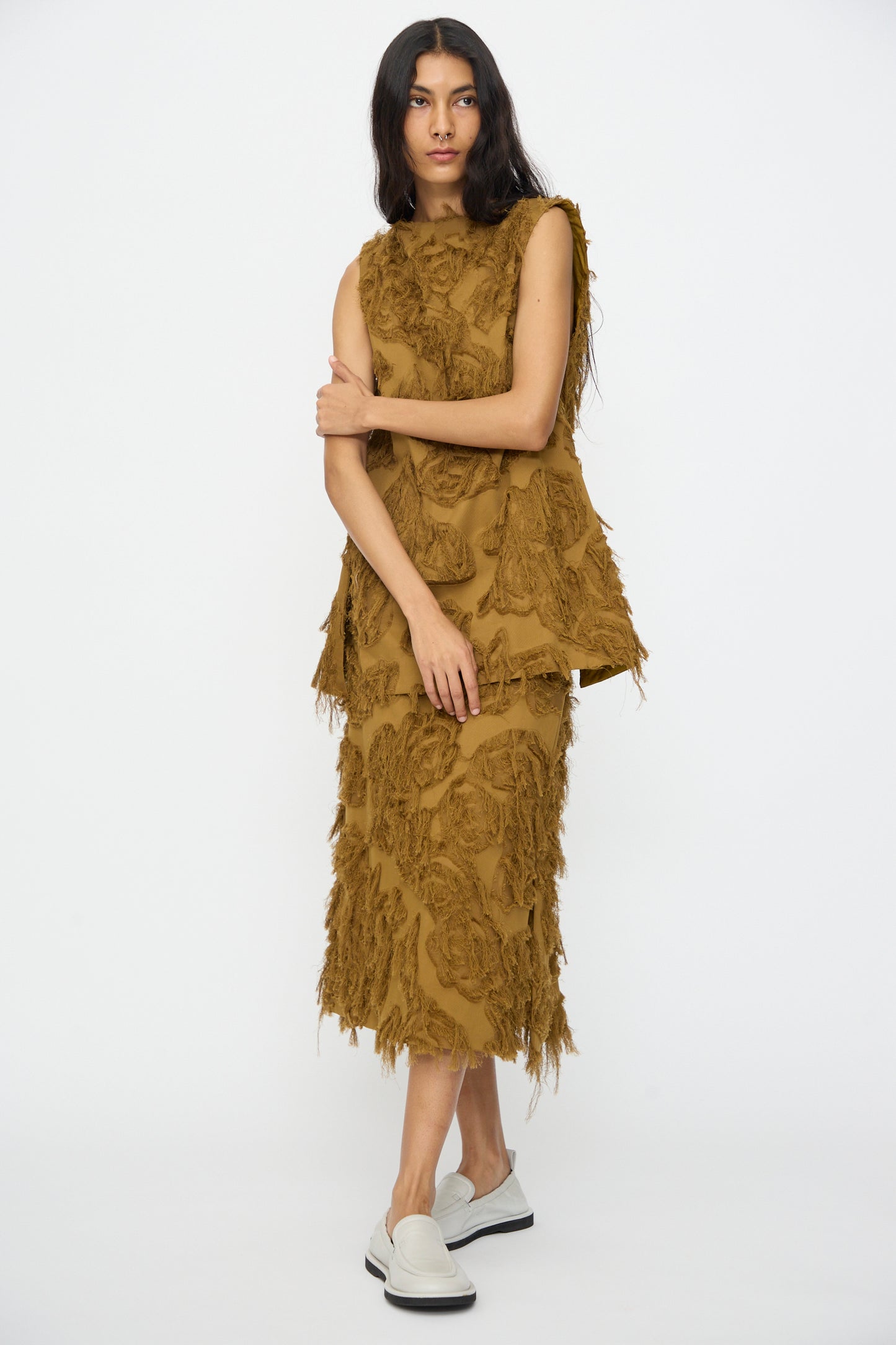 A woman stands against a white background wearing the Studio Nicholson Fringe Jacquard Mager Top in Mustard and a matching skirt with fringe, paired with white slip-on shoes.