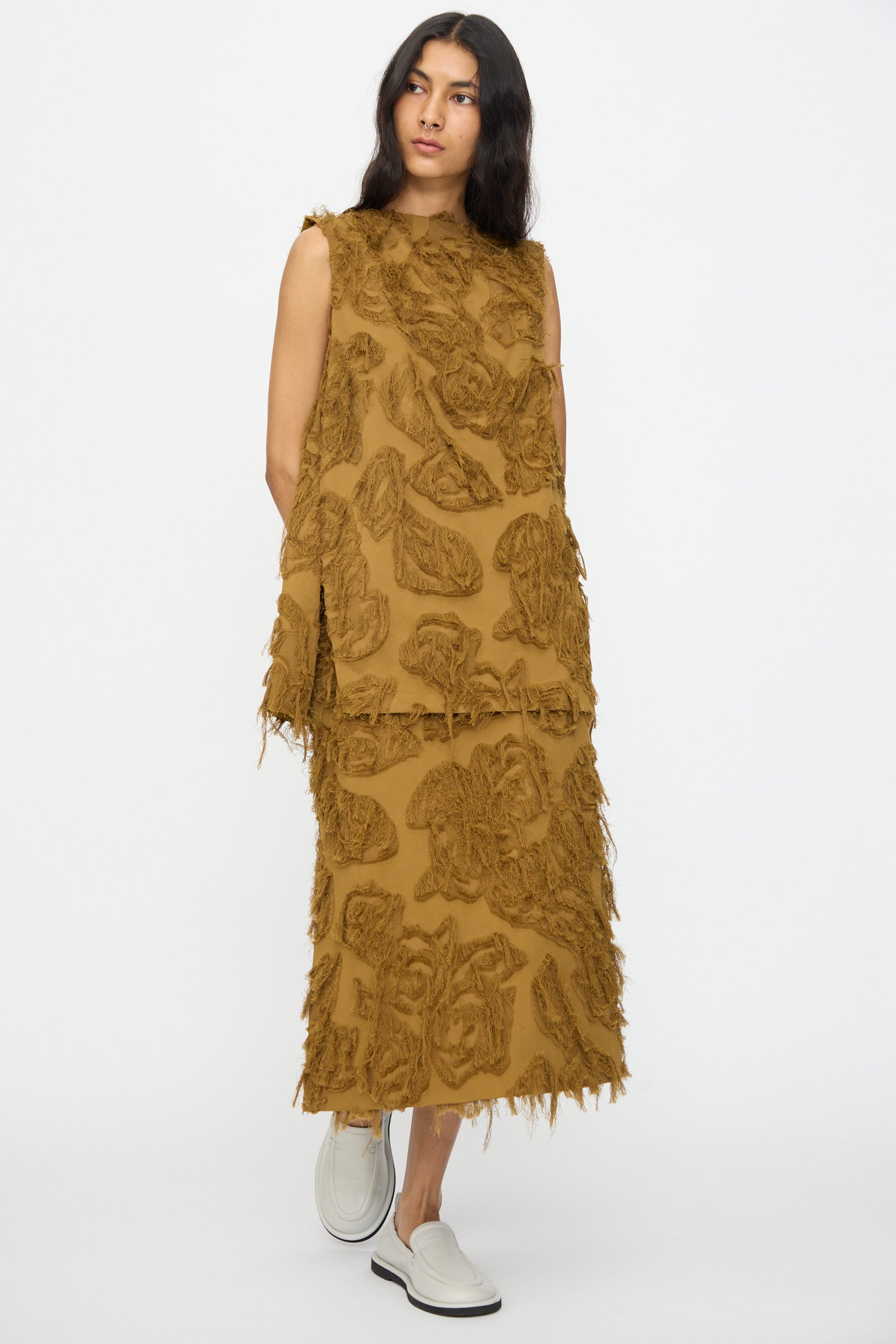 A woman wears a sleeveless brown cotton top and the Studio Nicholson Fringe Jacquard Roeper Skirt in Mustard with floral patterns, paired with white slip-on shoes, standing against a plain white background.