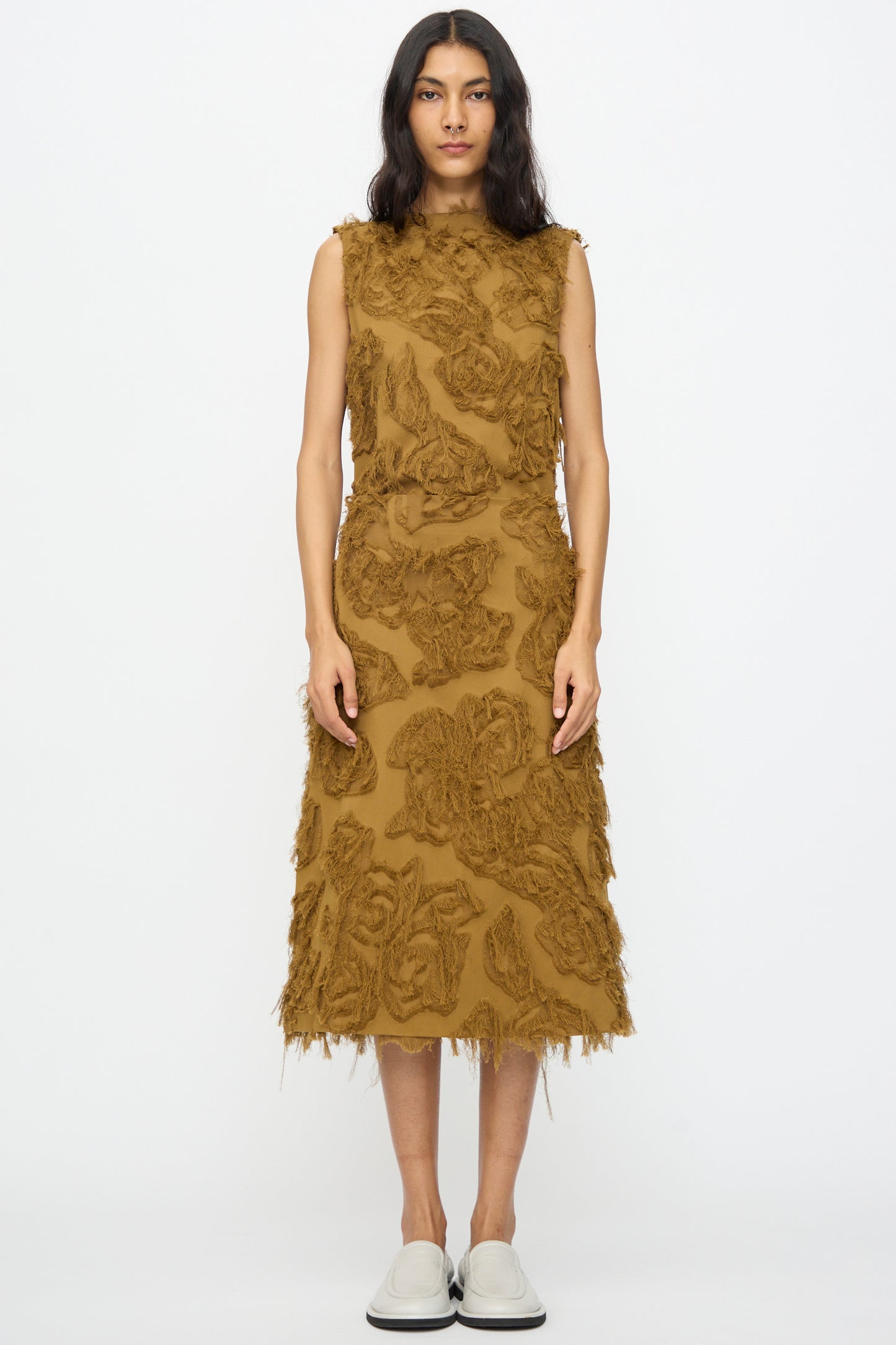 A woman stands facing forward wearing the Studio Nicholson Fringe Jacquard Roeper Skirt in mustard with a floral pattern and white slip-on shoes against a plain white background.