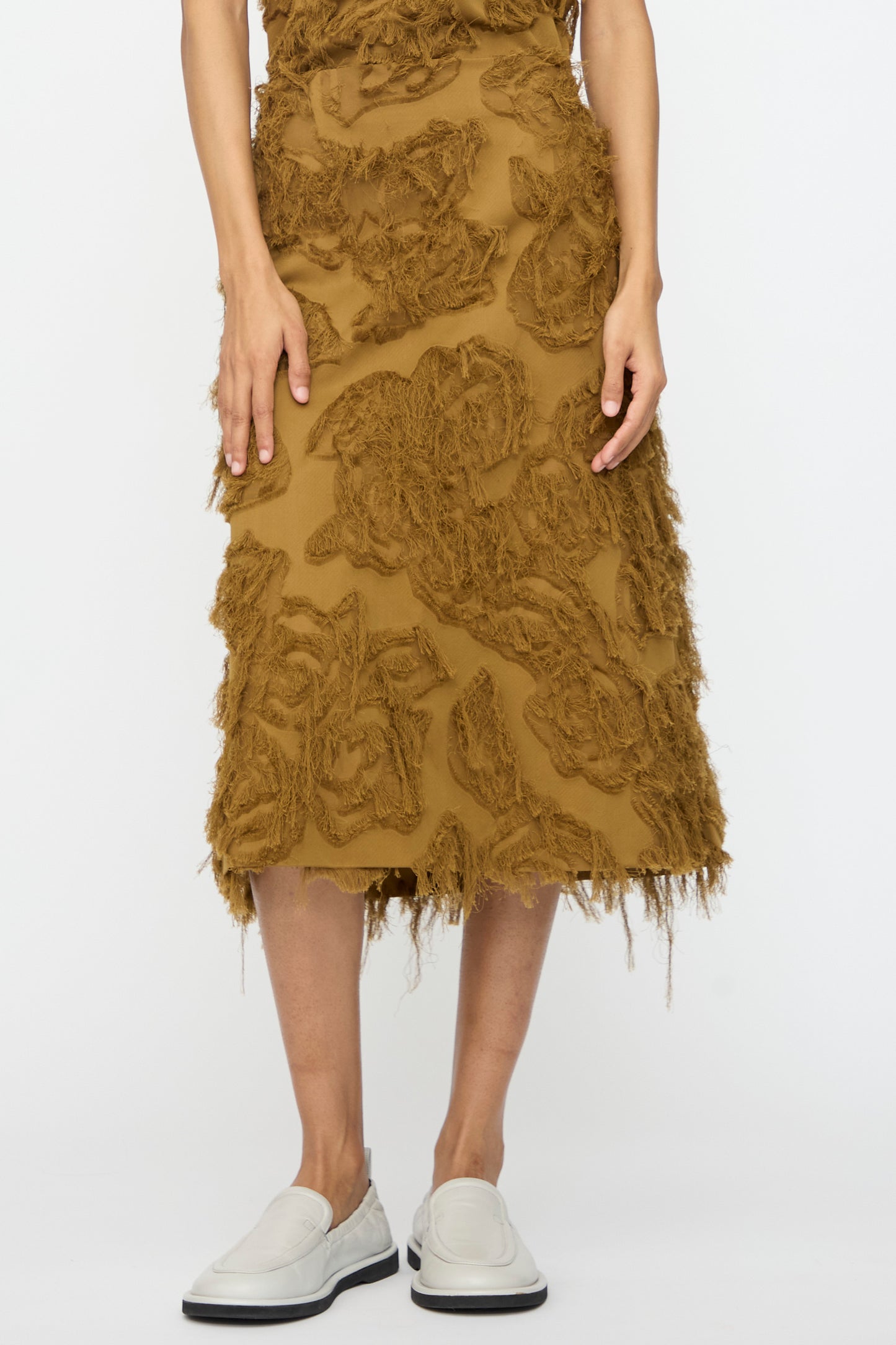 A person wears the Studio Nicholson Fringe Jacquard Roeper Skirt in mustard and white slip-on shoes, standing against a plain white background.