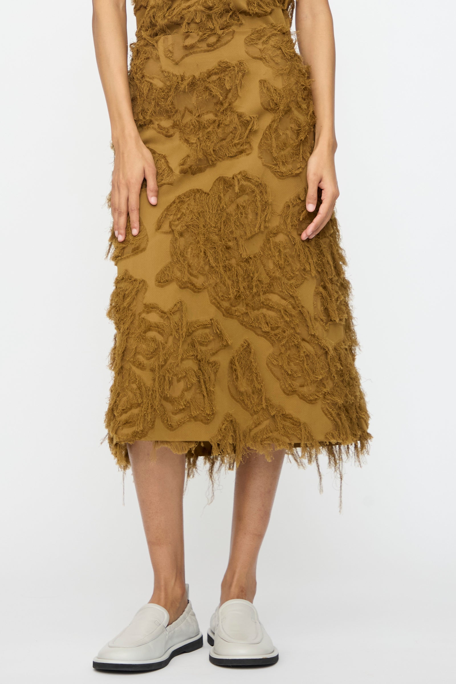 A person wears the Studio Nicholson Fringe Jacquard Roeper Skirt in mustard and white slip-on shoes, standing against a plain white background.