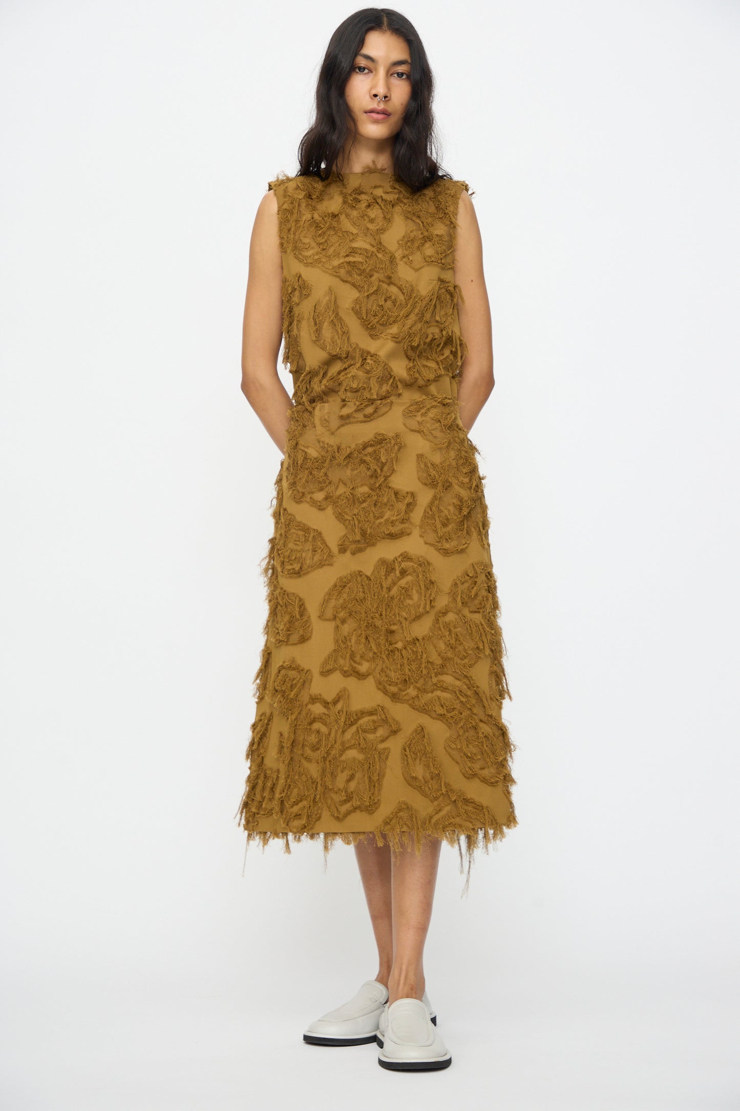 A woman stands against a white background wearing the Studio Nicholson Fringe Jacquard Roeper Skirt in mustard, featuring textured floral patterns and fringe details, paired with white slip-on shoes.