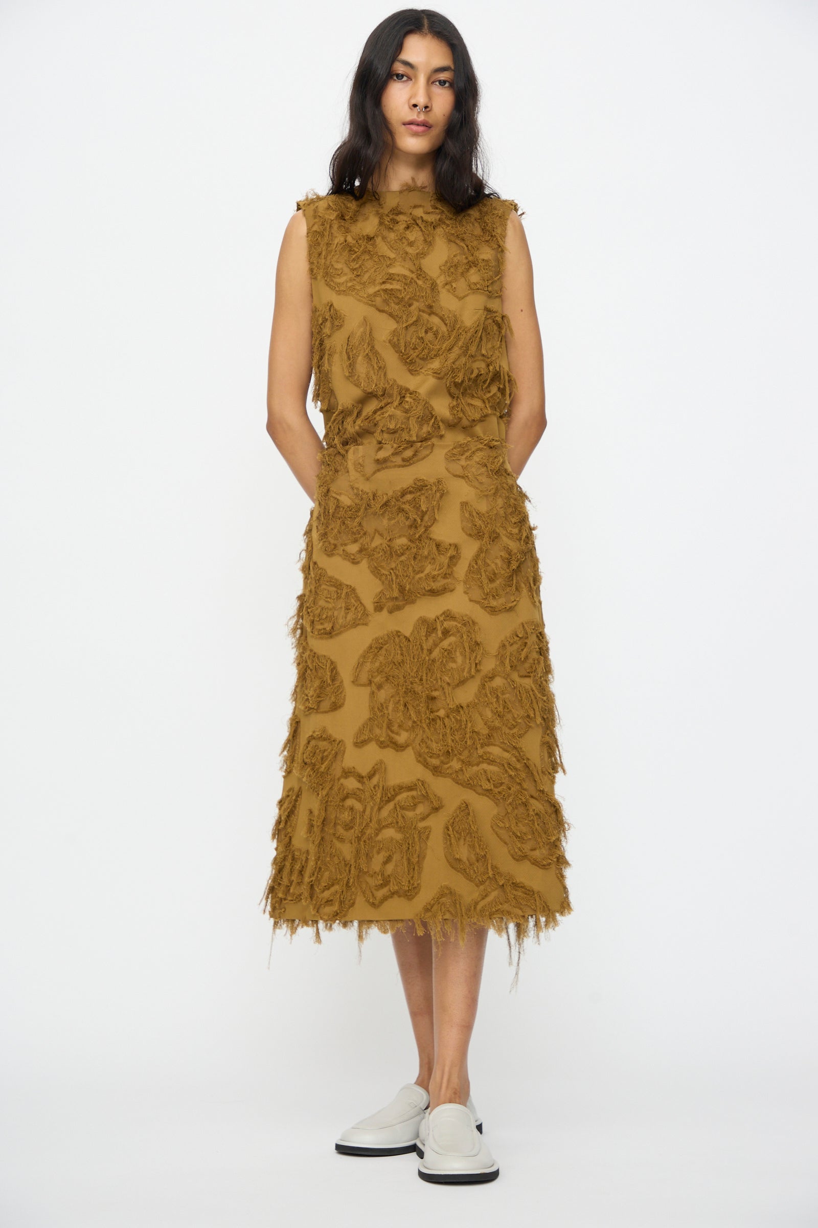 A woman stands against a white background wearing the Studio Nicholson Fringe Jacquard Roeper Skirt in mustard, featuring textured floral patterns and fringe details, paired with white slip-on shoes.