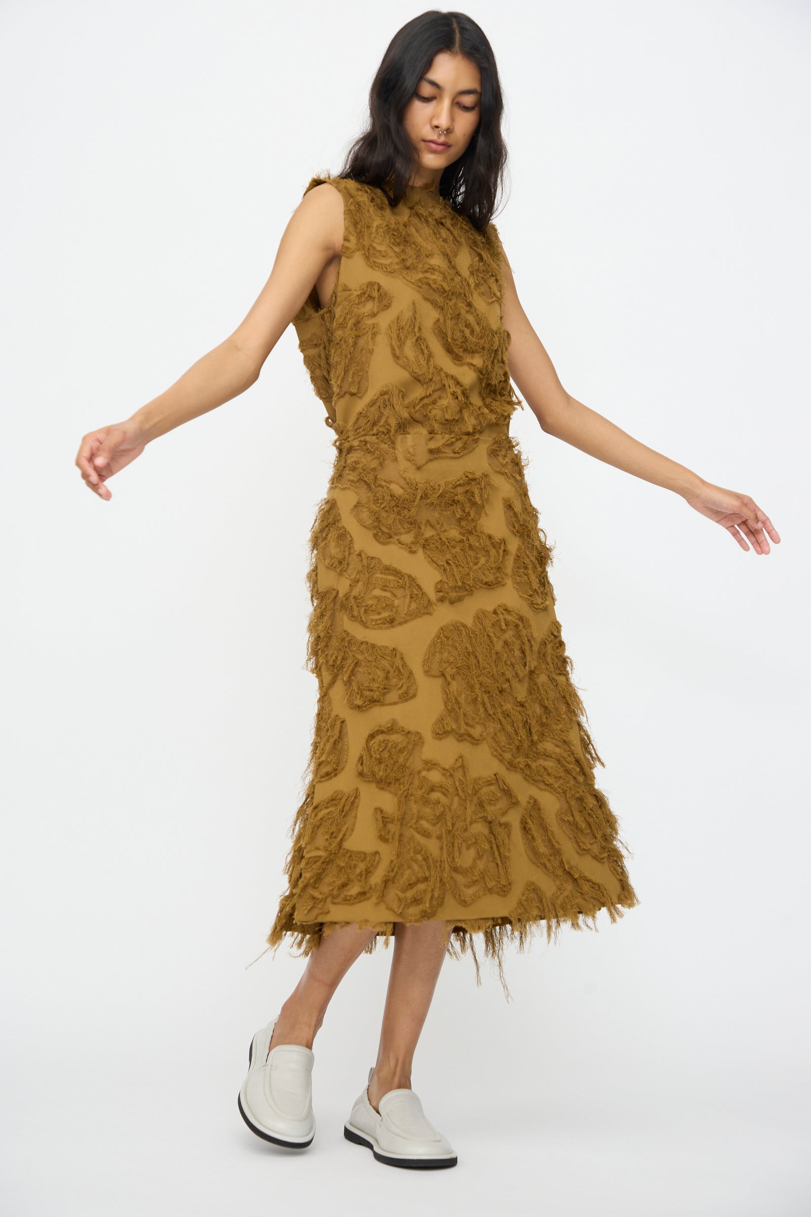 A woman wears the Studio Nicholson Fringe Jacquard Roeper Skirt in Mustard, styled with white slip-on shoes, standing against a plain white background.