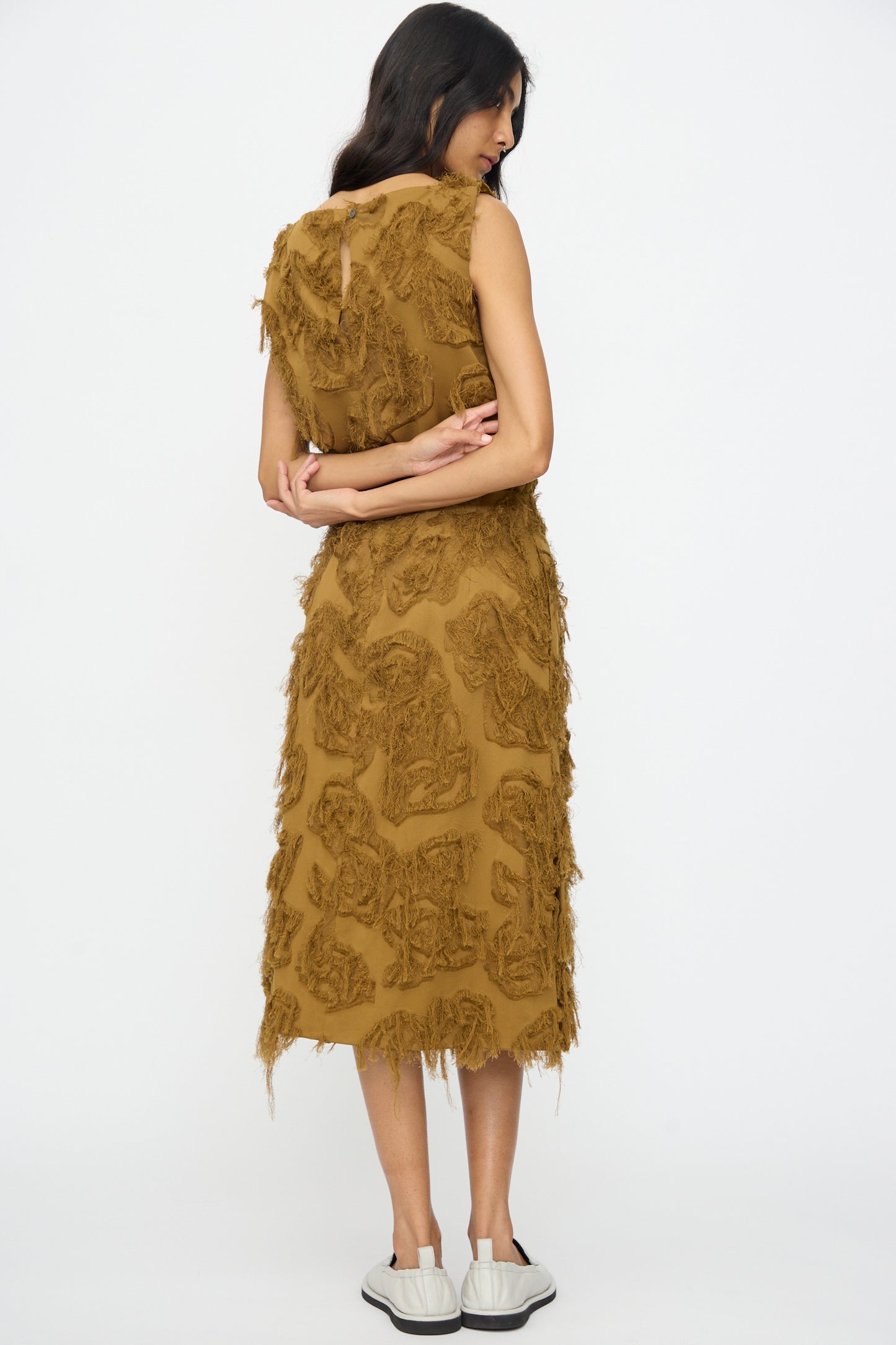 A woman faces away from the camera against a white background, arms crossed, wearing the Studio Nicholson Fringe Jacquard Roeper Skirt in Mustard and white sneakers.