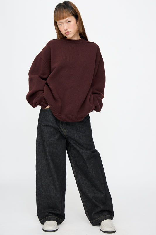 A person wearing an oversized, dark maroon Studio Nicholson Hemyl Guernsey Jumper in Grape with drop shoulders, loose dark denim jeans, and platform shoes.