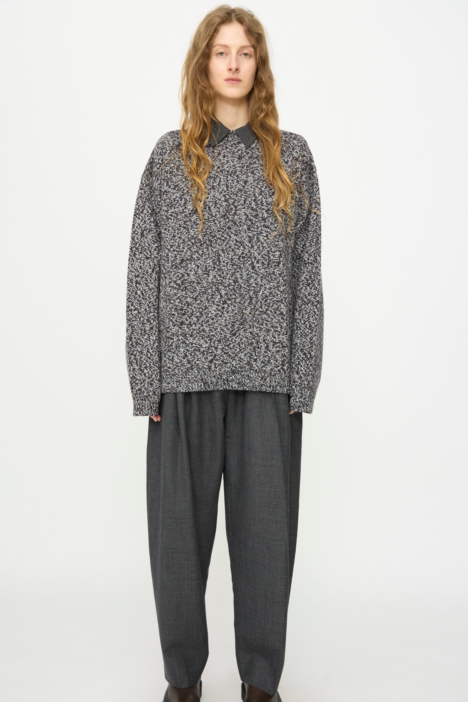 A person stands against a white background wearing Studio Nicholson’s Hemyl Guernsey Jumper in Navy Mouline over a collared shirt, paired with loose dark gray trousers and dark shoes.