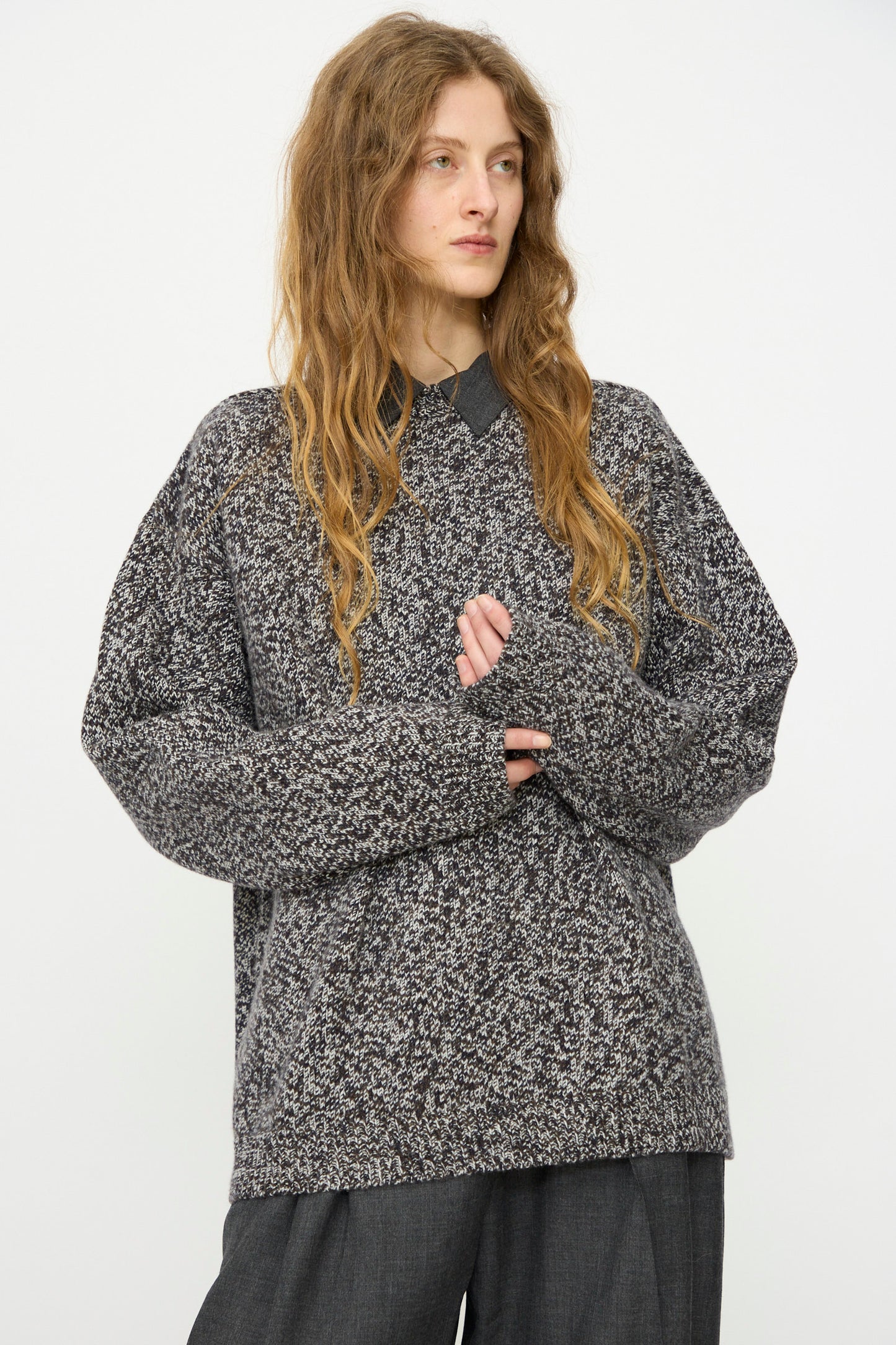 Person with long wavy hair wears the Studio Nicholson Hemyl Guernsey Jumper in Navy Mouline, made in London, layered over a collared shirt and dark trousers, standing against a plain white background.