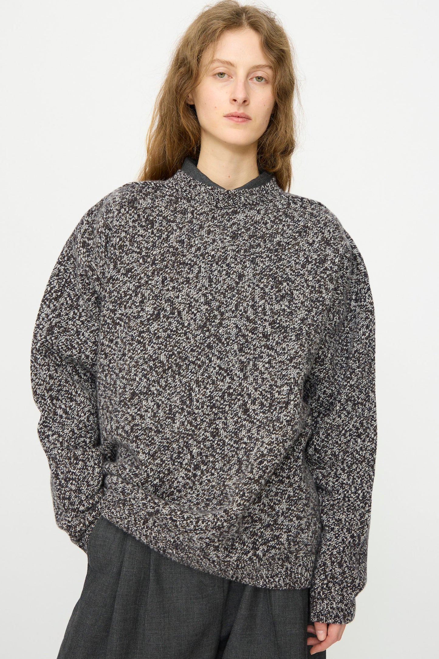 A person with long, wavy hair wears the Studio Nicholson Hemyl Guernsey Jumper in Navy Mouline and dark pants, standing against a plain white background.