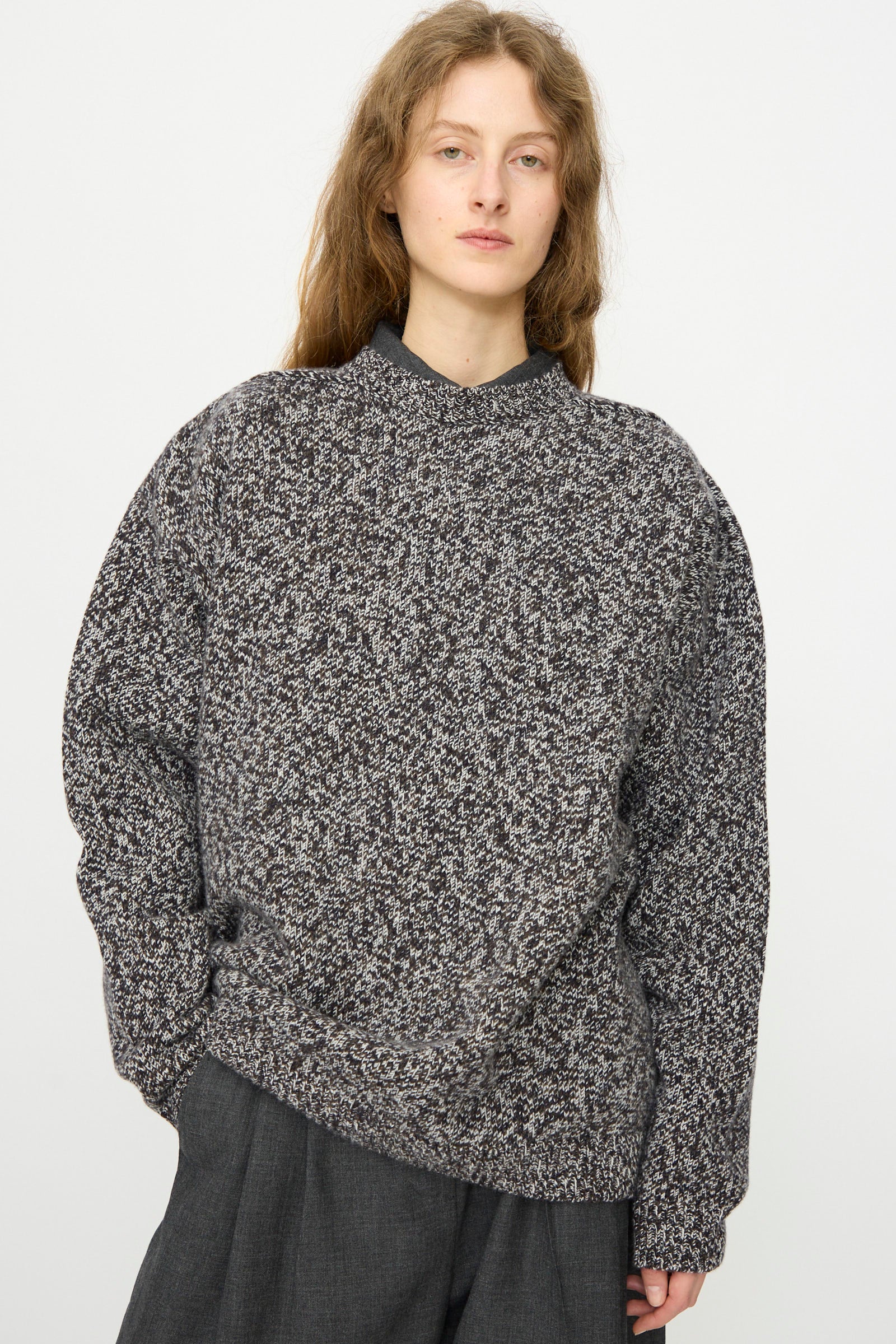 A person with long, wavy hair wears the Studio Nicholson Hemyl Guernsey Jumper in Navy Mouline and dark pants, standing against a plain white background.