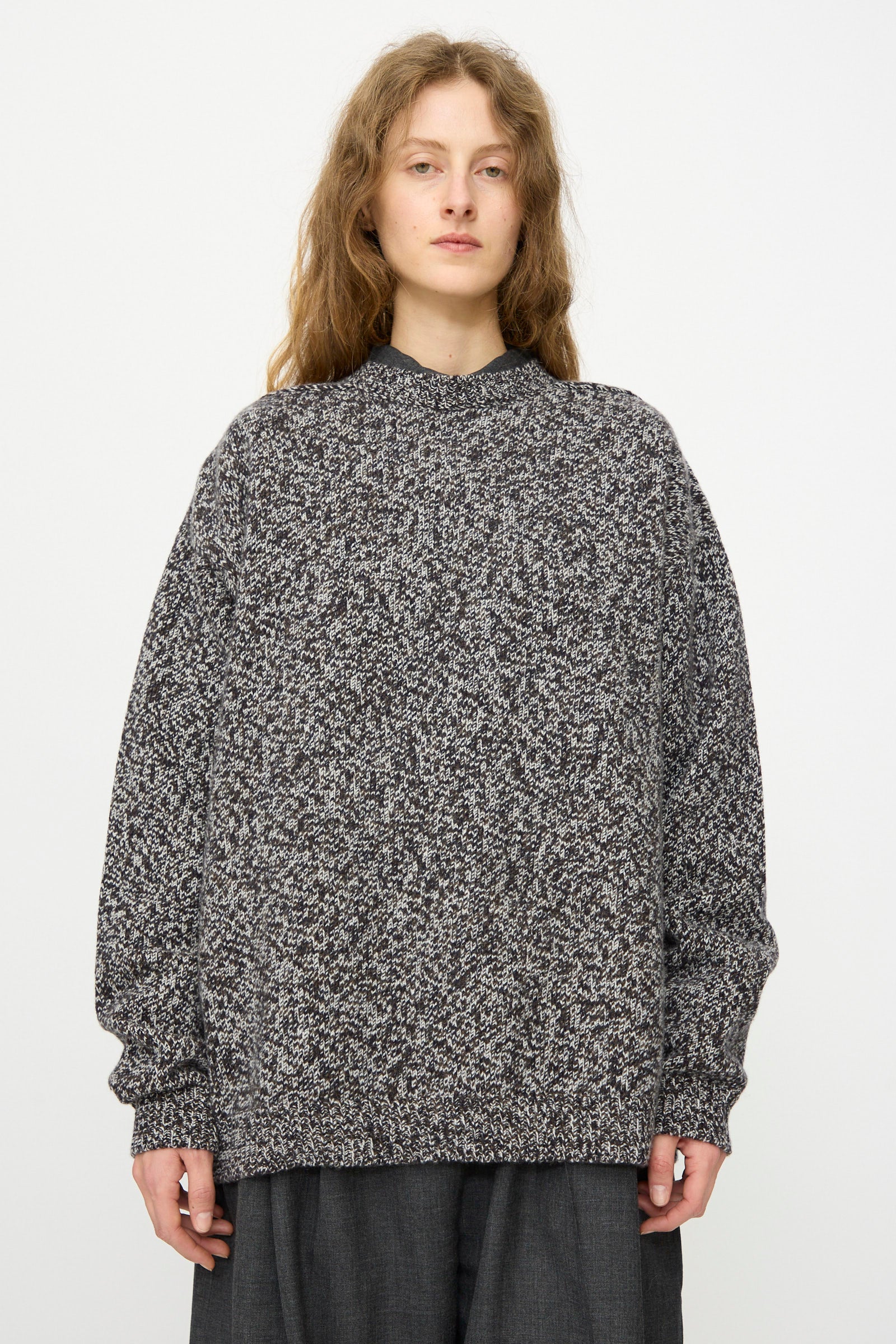 A person with long, wavy light brown hair wears the Studio Nicholson Hemyl Guernsey Jumper in Navy Mouline and dark pants, standing against a plain light background.