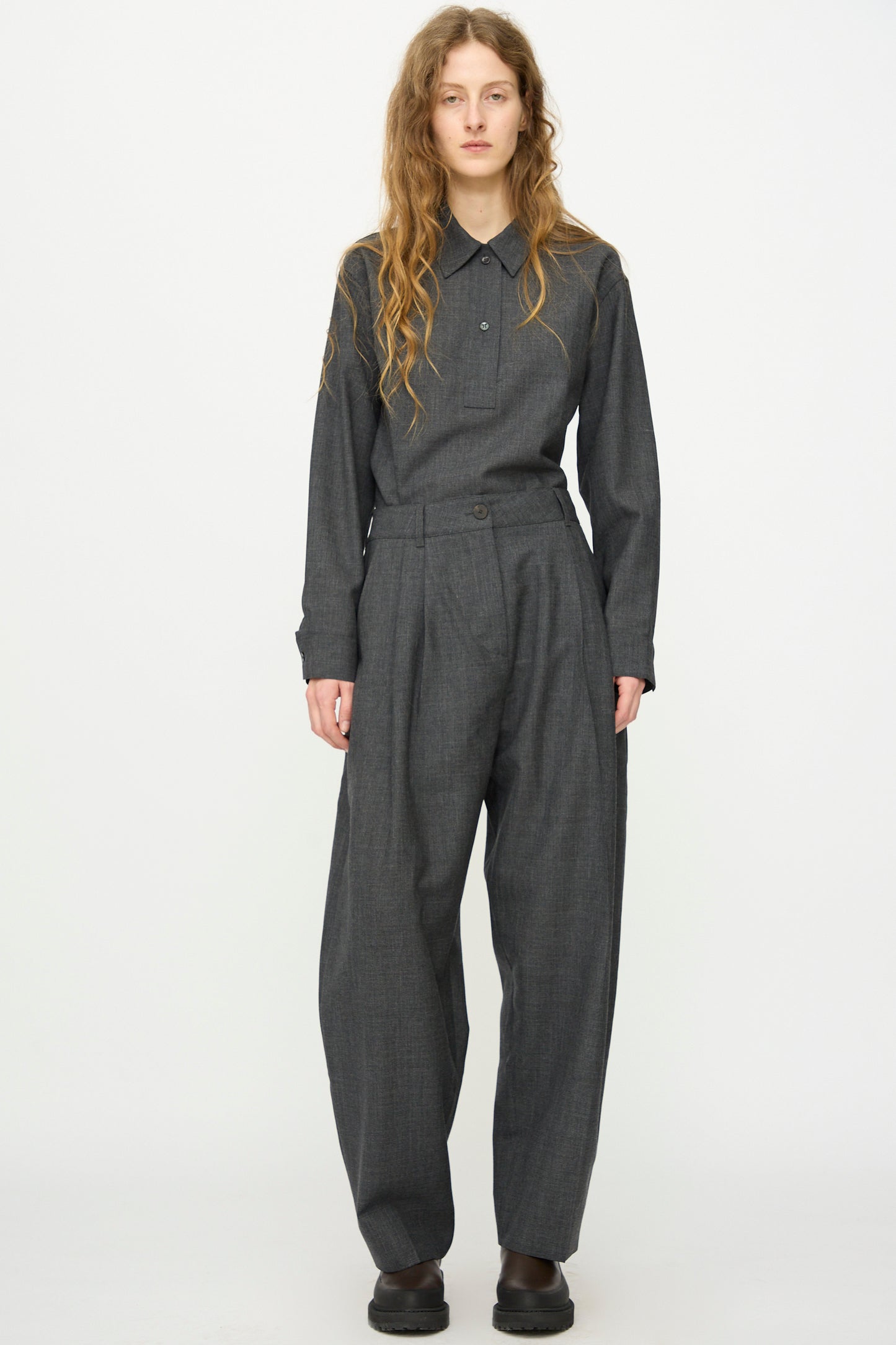 A person with long wavy hair wears a charcoal grey button-up shirt tucked into Studio Nicholson's Heritage Wool Acuna Pant in Slate Melange, paired with black shoes, standing against a plain white background.