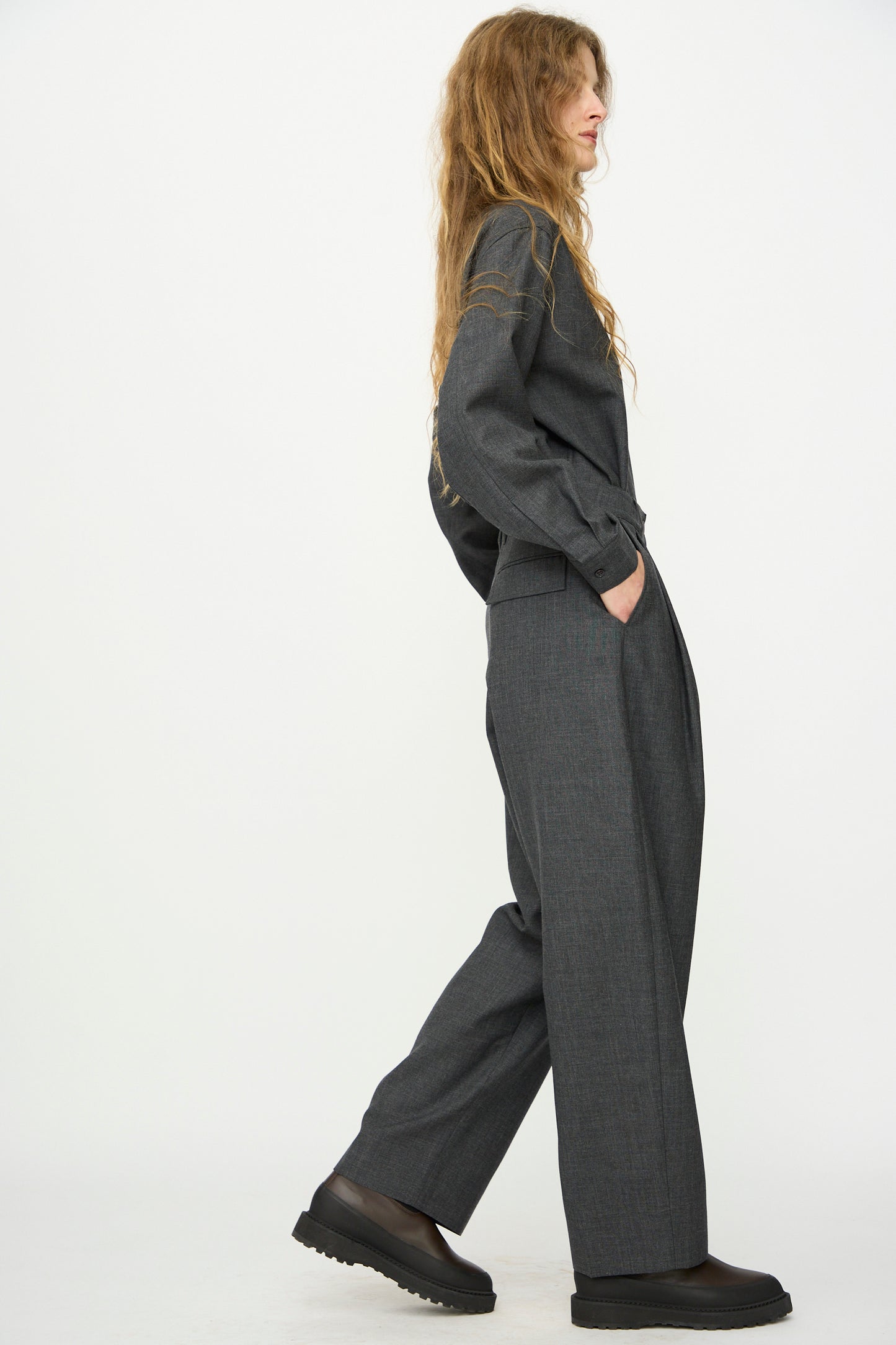 A person with long wavy hair stands in profile, hands in pockets, wearing Studio Nicholson's Heritage Wool Acuna Pant in Slate Melange and a loose-fitting dark gray suit with black shoes against a plain white background.