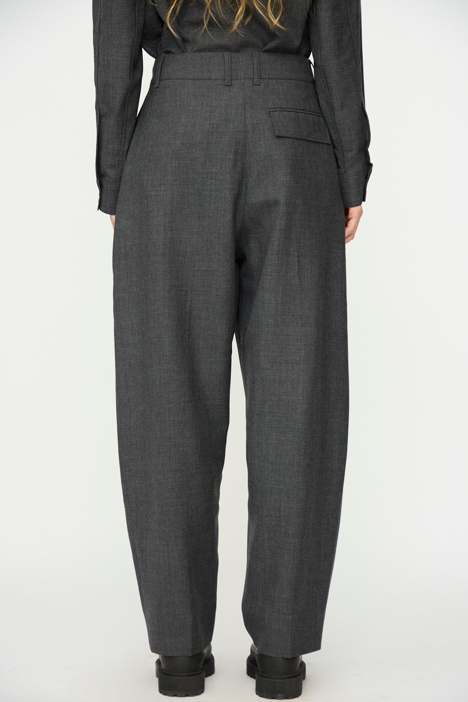 A person wearing Studio Nicholson's Heritage Wool Acuna Pant in Slate Melange and black shoes stands with their back to the camera against a plain background.