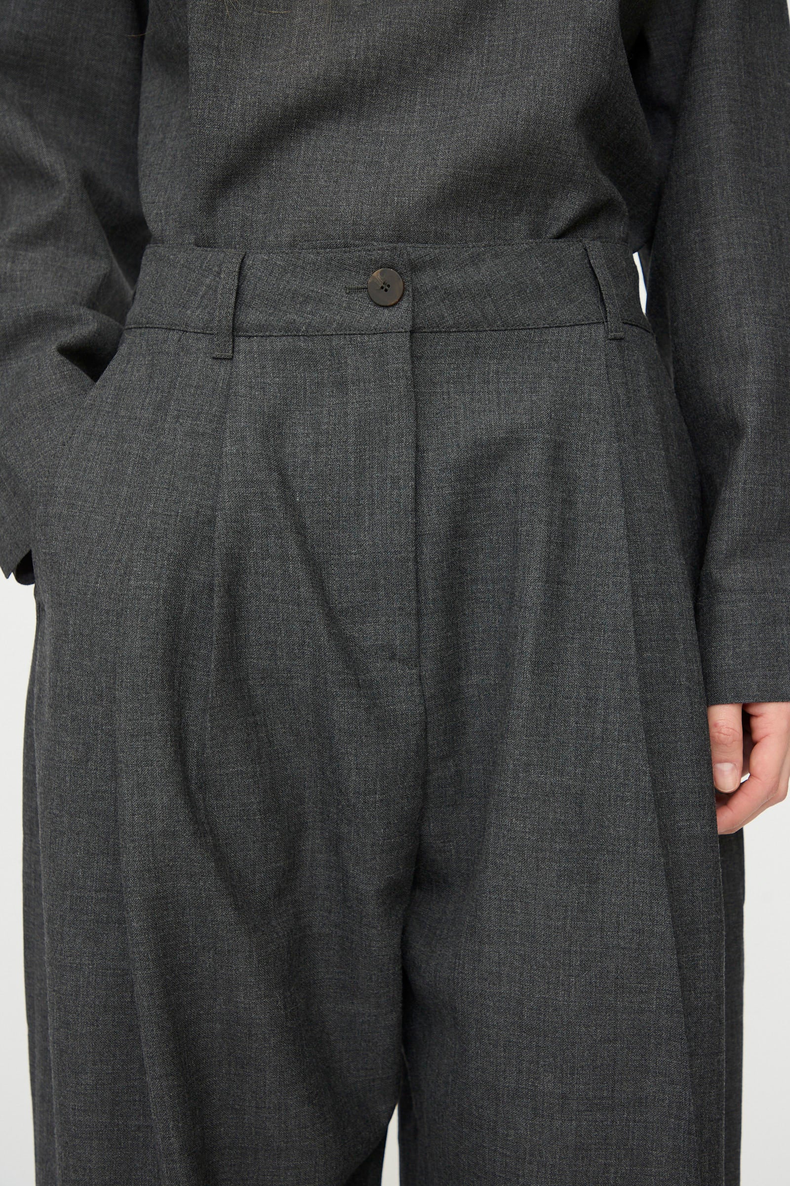 Close-up of a person wearing Studio Nicholson's Heritage Wool Acuna Pant in Slate Melange, featuring a high-waisted pleated design and buttoned waistband.