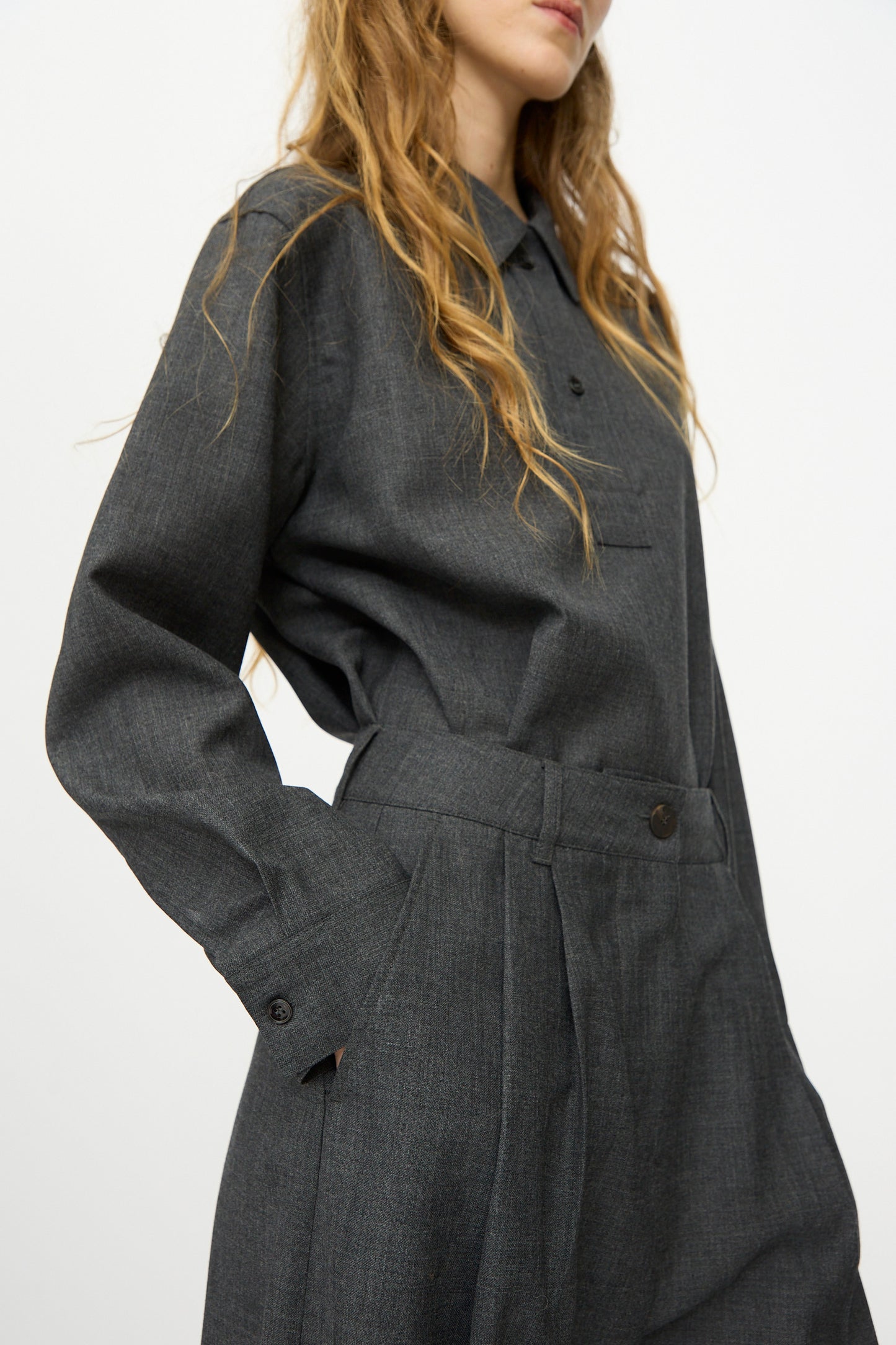 A woman with long wavy hair wears a dark gray button-up shirt and Studio Nicholson's Heritage Wool Acuna Pant in Slate Melange, standing against a plain white background.