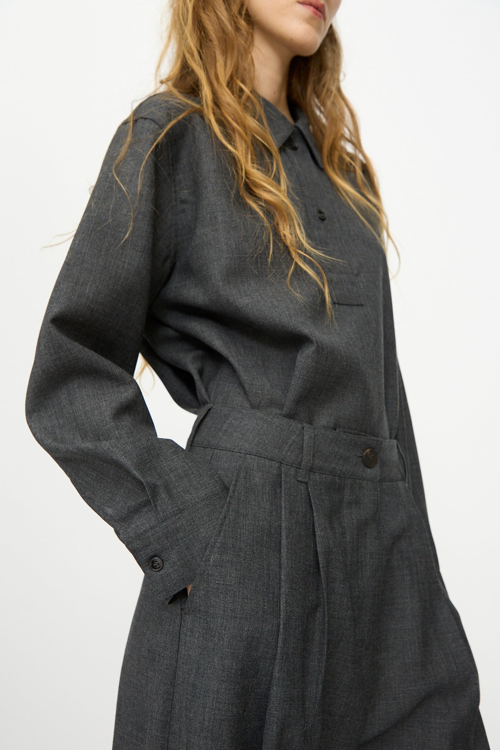 A woman with long wavy hair wears a dark gray button-up shirt and Studio Nicholson's Heritage Wool Acuna Pant in Slate Melange, standing against a plain white background.