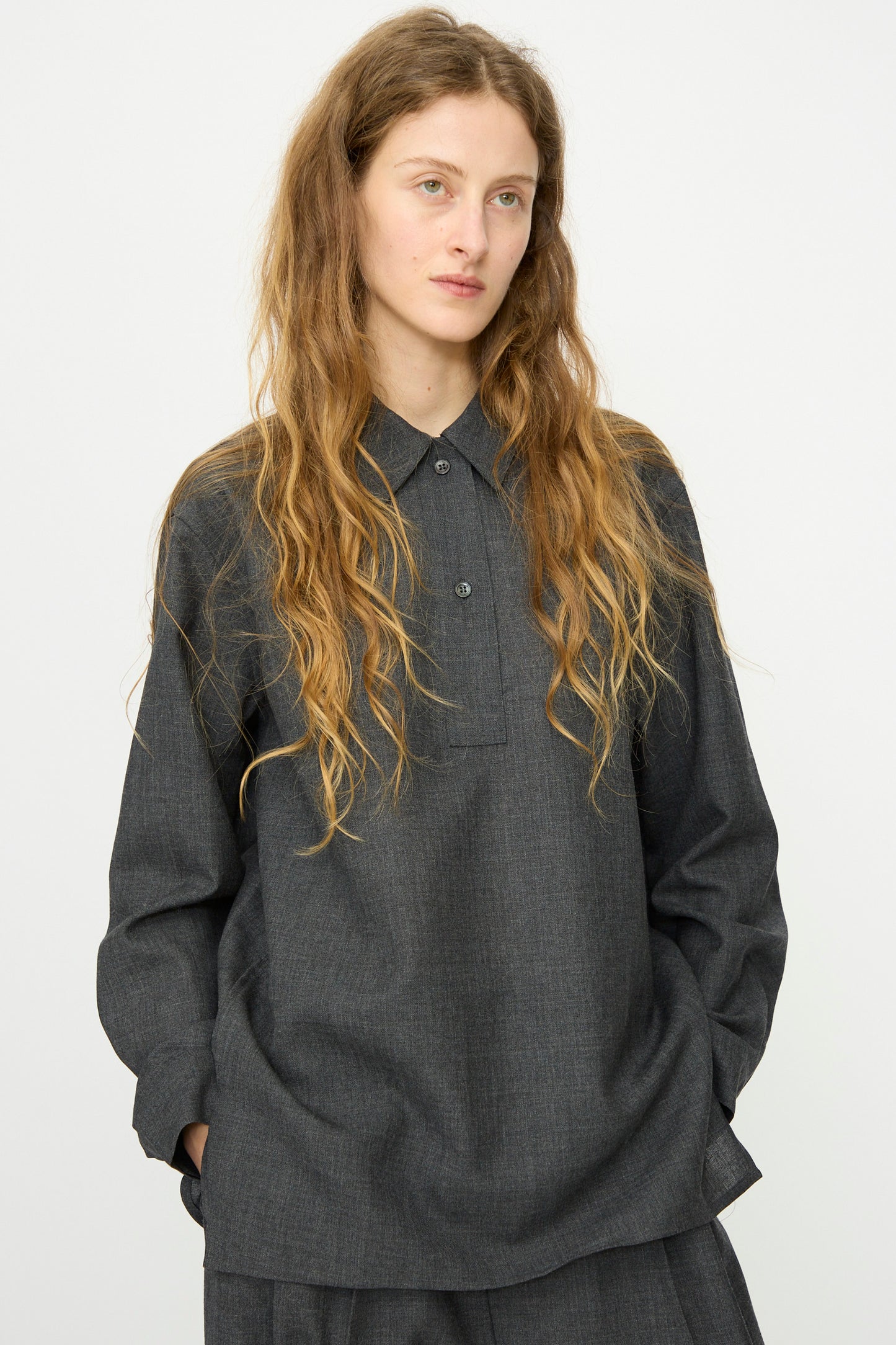 A person with long, wavy hair stands against a plain light background, hands in pockets, wearing Studio Nicholson's relaxed-fit Heritage Wool Pinole Shirt in Slate Melange and matching pants.