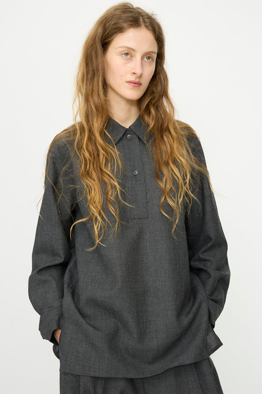 A person with long, wavy hair stands against a plain light background, hands in pockets, wearing Studio Nicholson's relaxed-fit Heritage Wool Pinole Shirt in Slate Melange and matching pants.