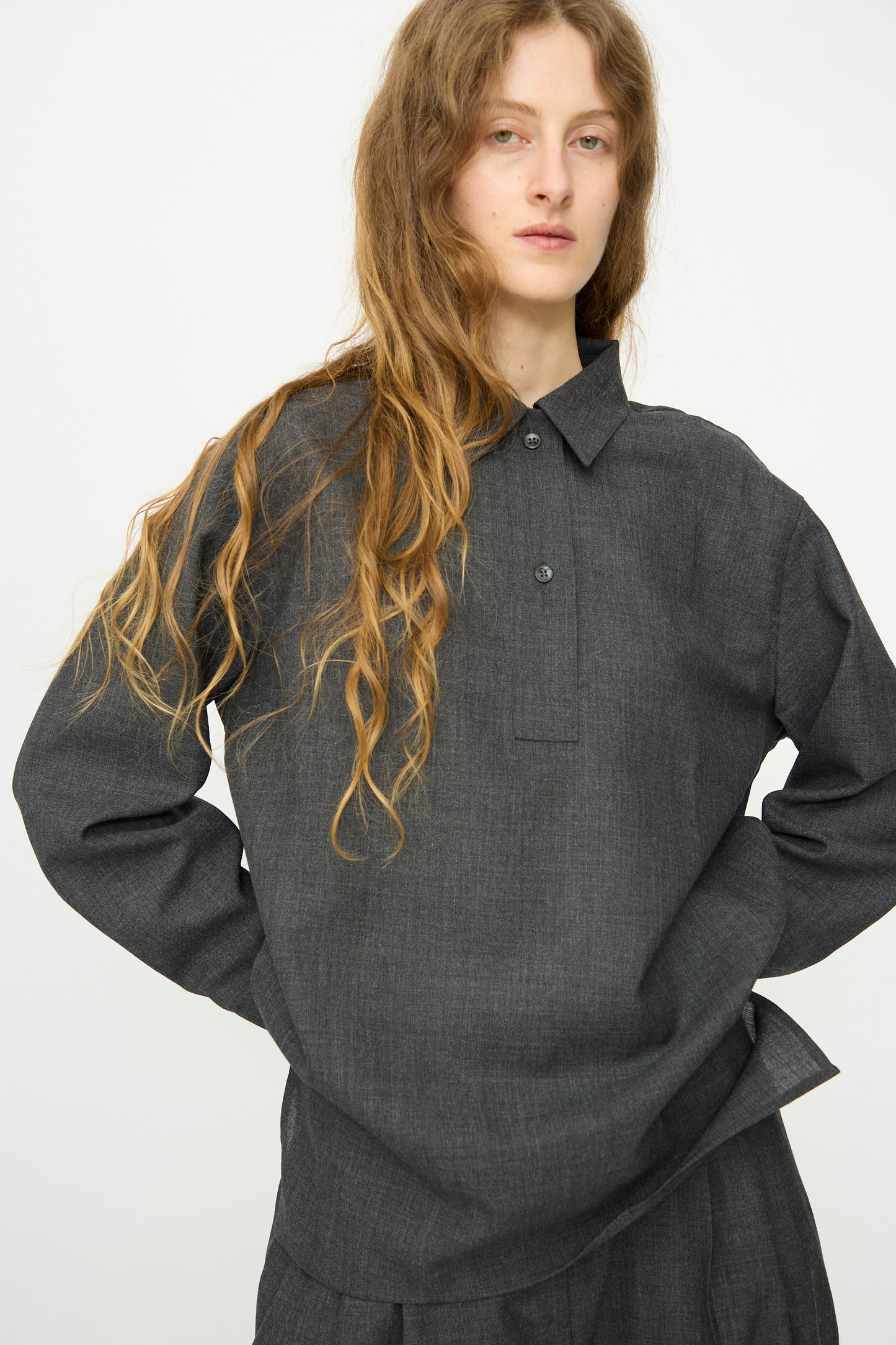 A person with long, wavy hair wears a relaxed fit Studio Nicholson Heritage Wool Pinole Shirt in Slate Melange, paired with matching pants, standing against a plain light background.
