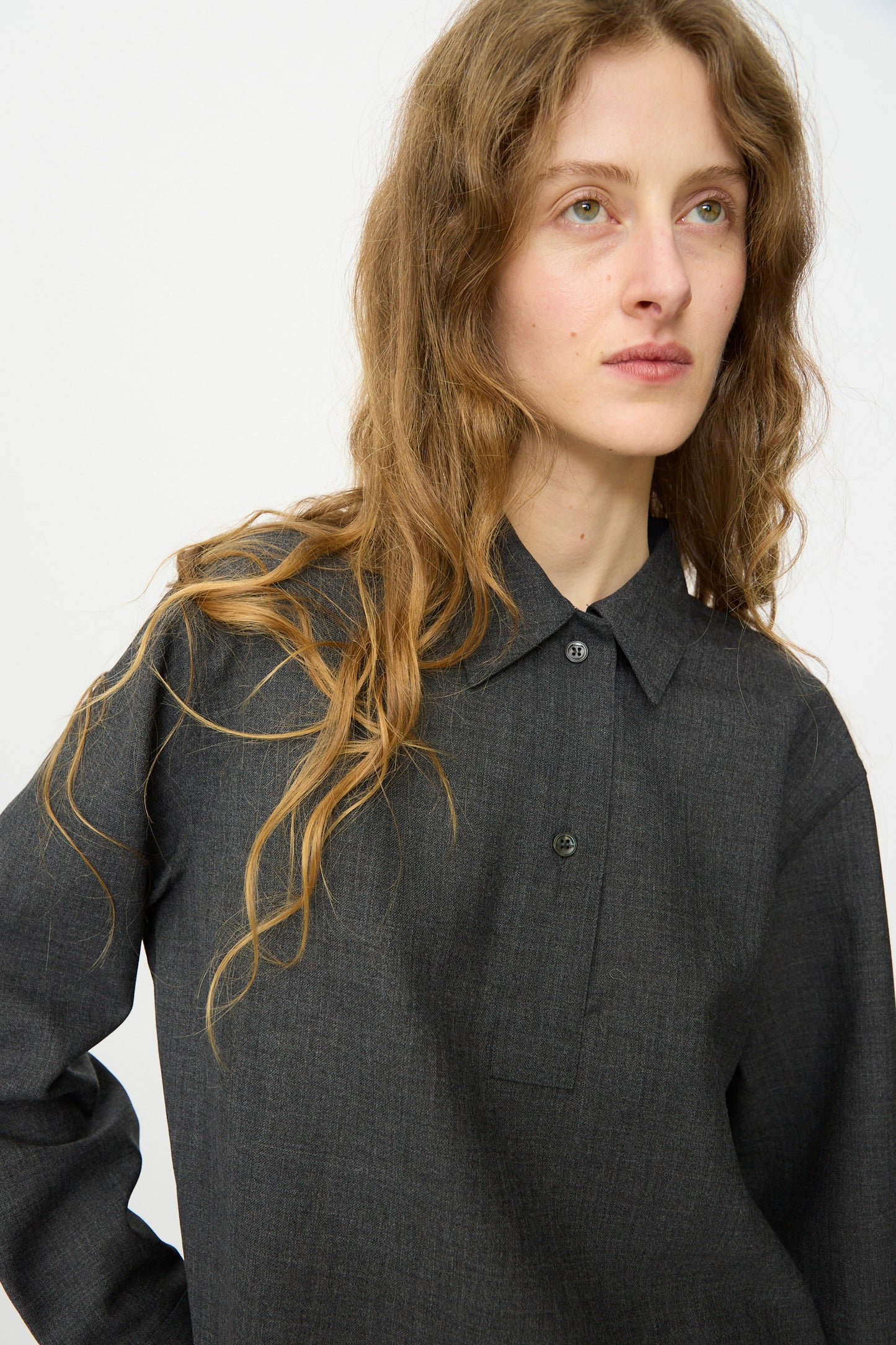 Someone with long wavy hair, wearing the Studio Nicholson Heritage Wool Pinole Shirt in Slate Melange, looks slightly to the side against a plain light background.