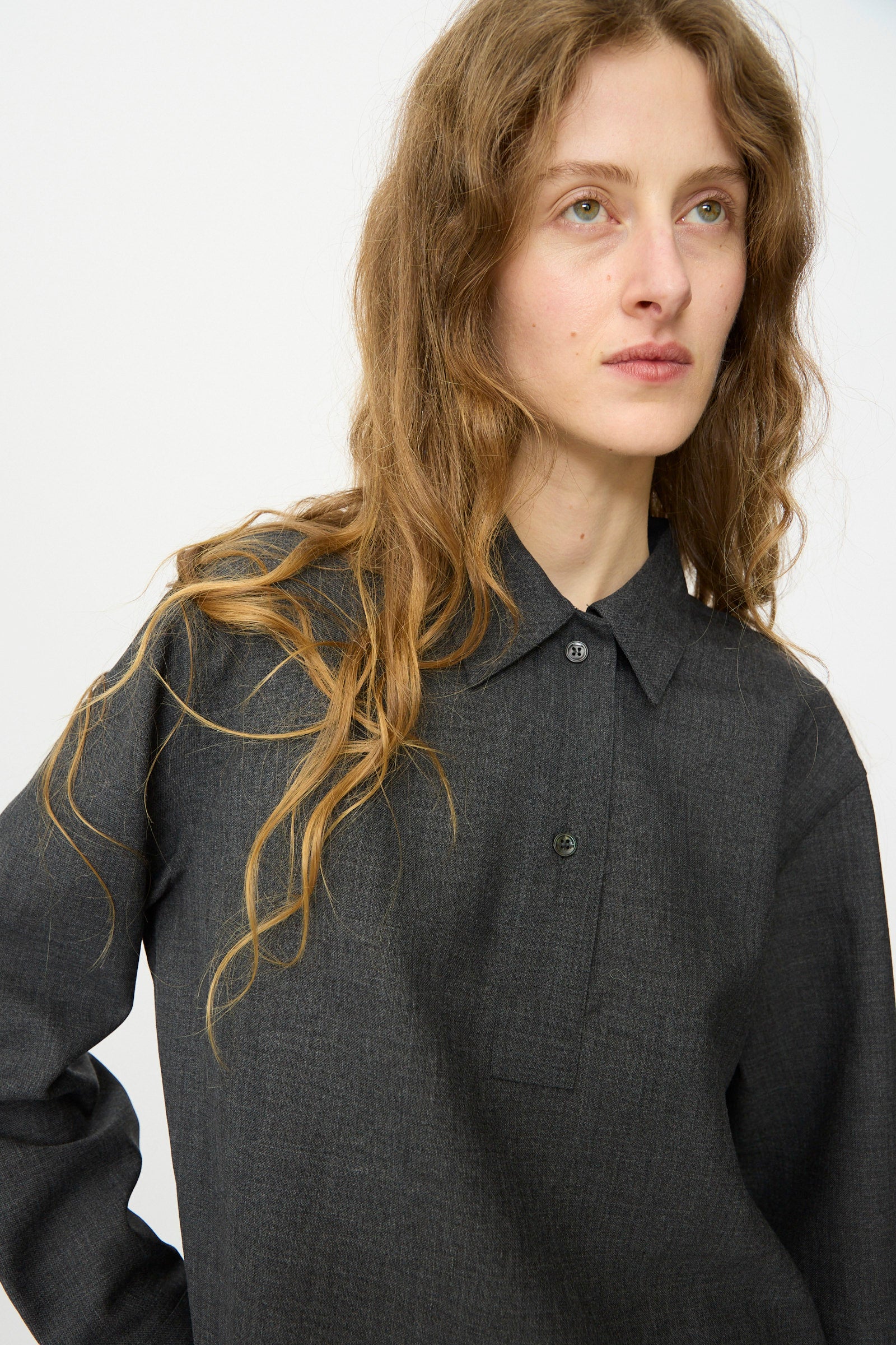 Someone with long wavy hair, wearing the Studio Nicholson Heritage Wool Pinole Shirt in Slate Melange, looks slightly to the side against a plain light background.