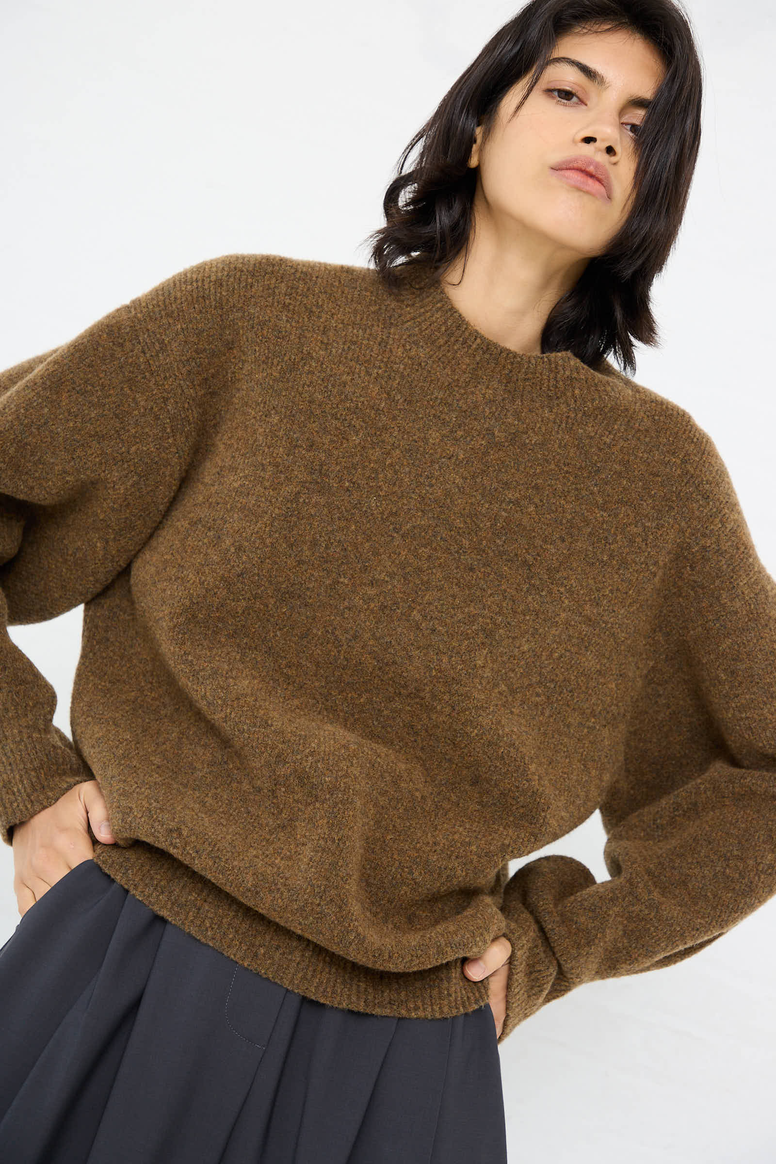 A person with shoulder-length hair wears a relaxed fit, High Loft Wool Sirio Crew Neck sweater in Nutmeg by Studio Nicholson and dark gray pants, standing against a white background with hands in pockets.