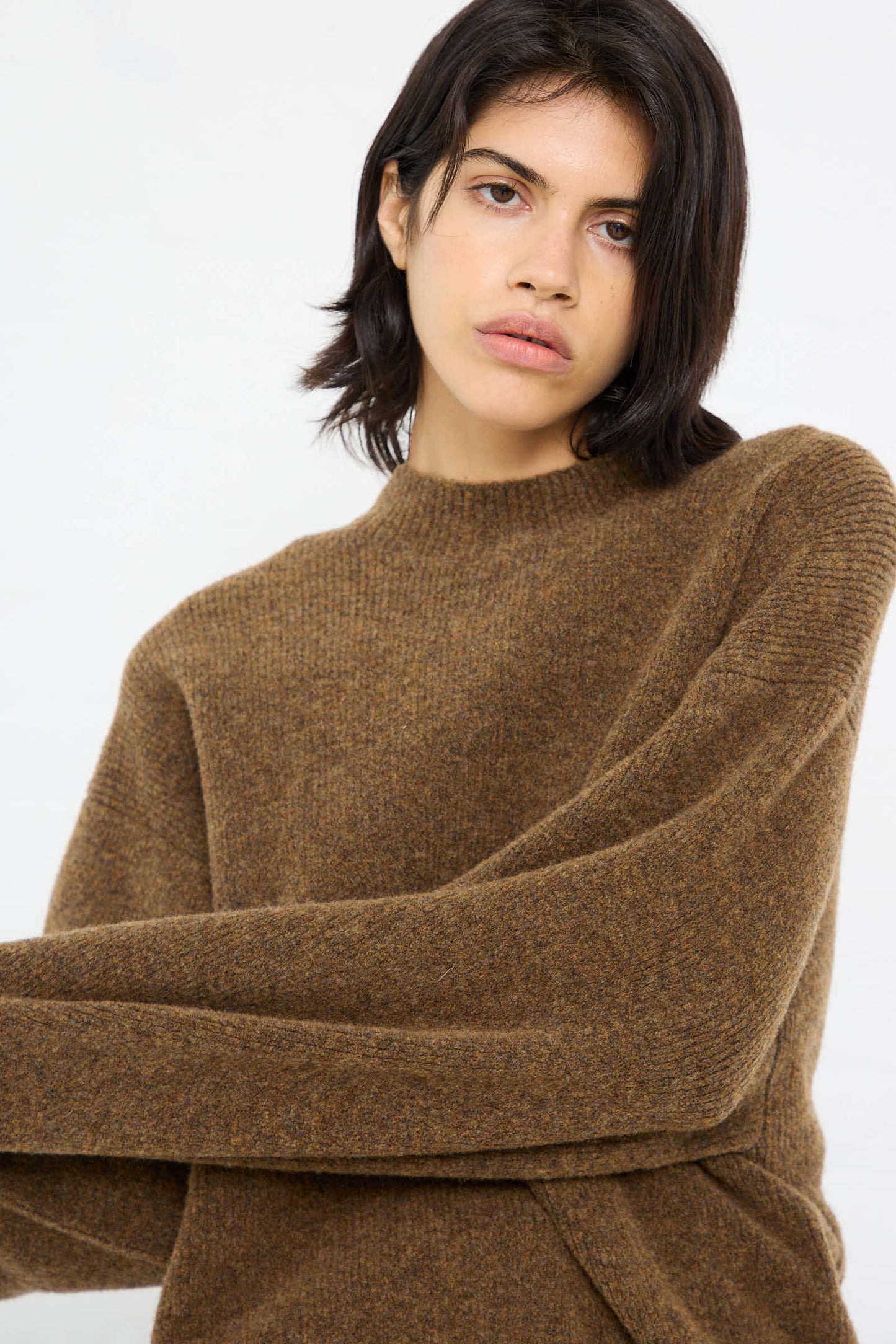 A person in a High Loft Wool Sirio Crew Neck in Nutmeg from Studio Nicholson, a brown long-sleeved sweater, stands against a plain background, looking slightly to the side. The relaxed fit and knit merino wool blend add both comfort and style.