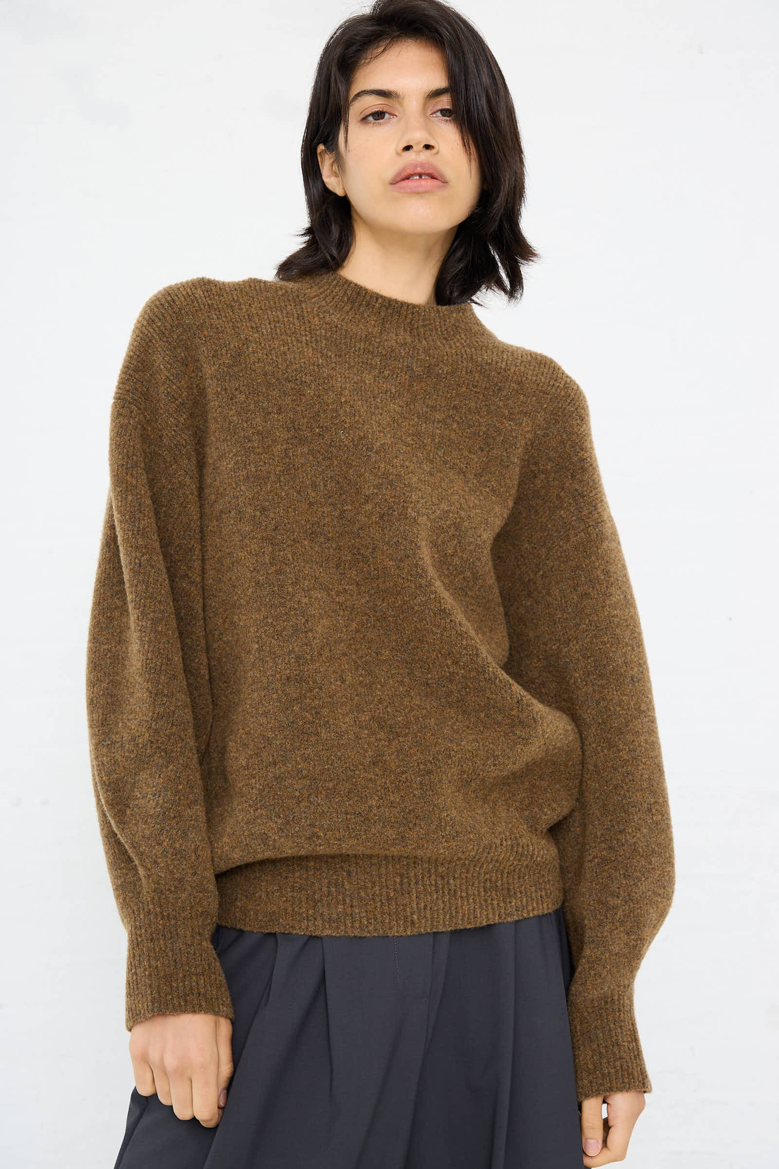 A person with shoulder-length dark hair is wearing a relaxed fit Studio Nicholson High Loft Wool Sirio Crew Neck in Nutmeg sweater and dark pants, standing against a plain white background.