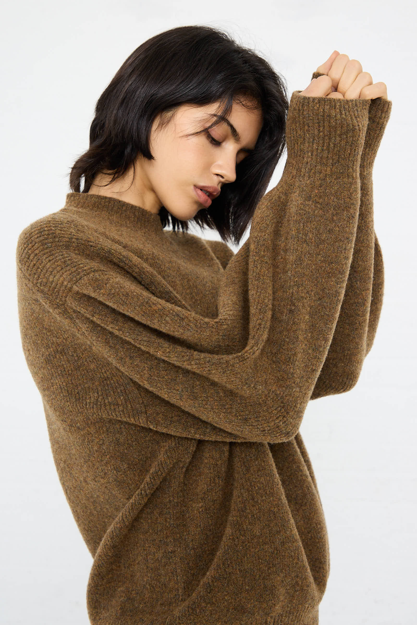 A woman wearing the Studio Nicholson High Loft Wool Sirio Crew Neck in Nutmeg, an oversized relaxed fit sweater, is tilting her head down with eyes closed and holding her hands together near her face against a plain white background.