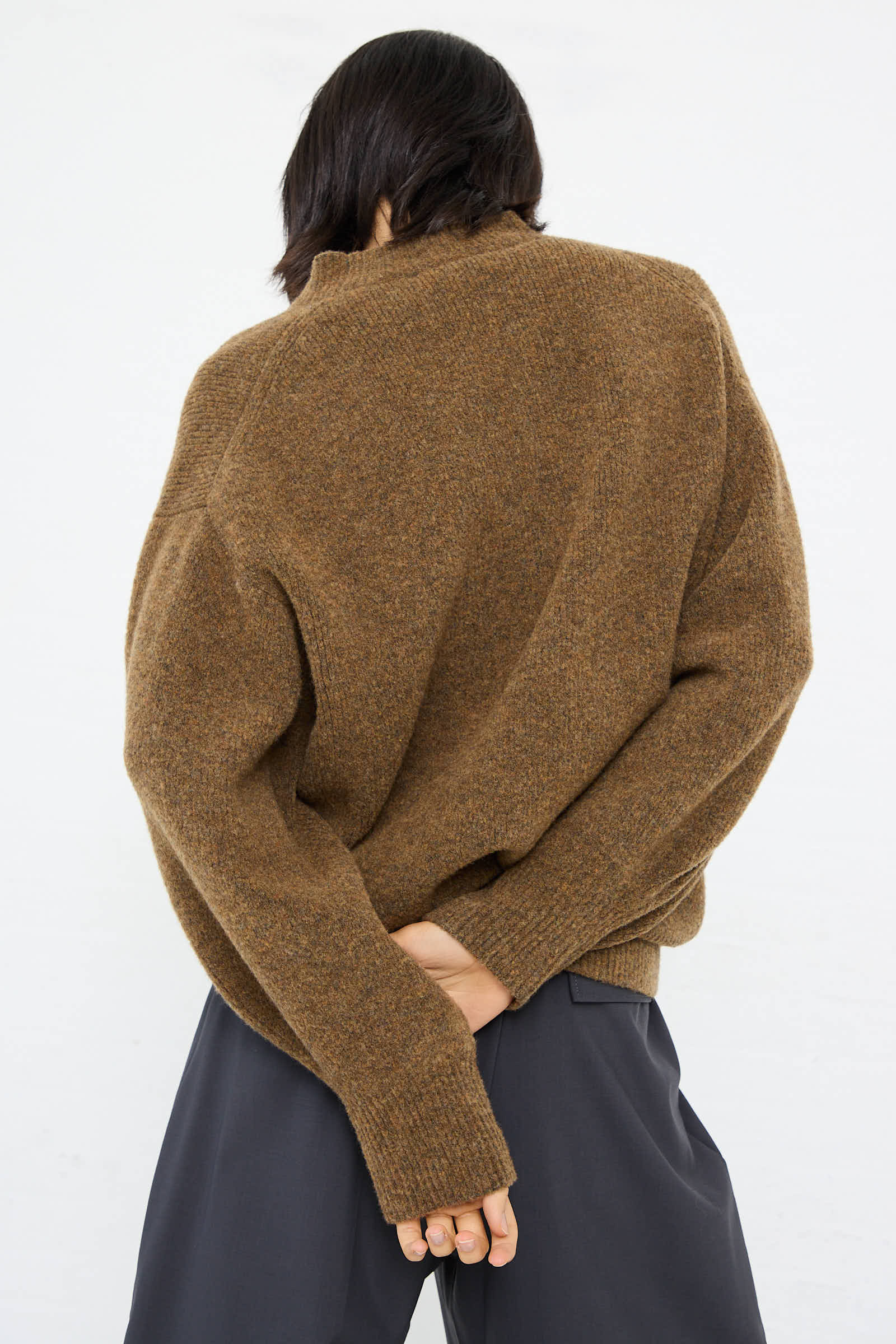 Person wearing an oversized, relaxed-fit High Loft Wool Sirio Crew Neck in Nutmeg by Studio Nicholson with dark pants, their back turned and left hand behind their back.