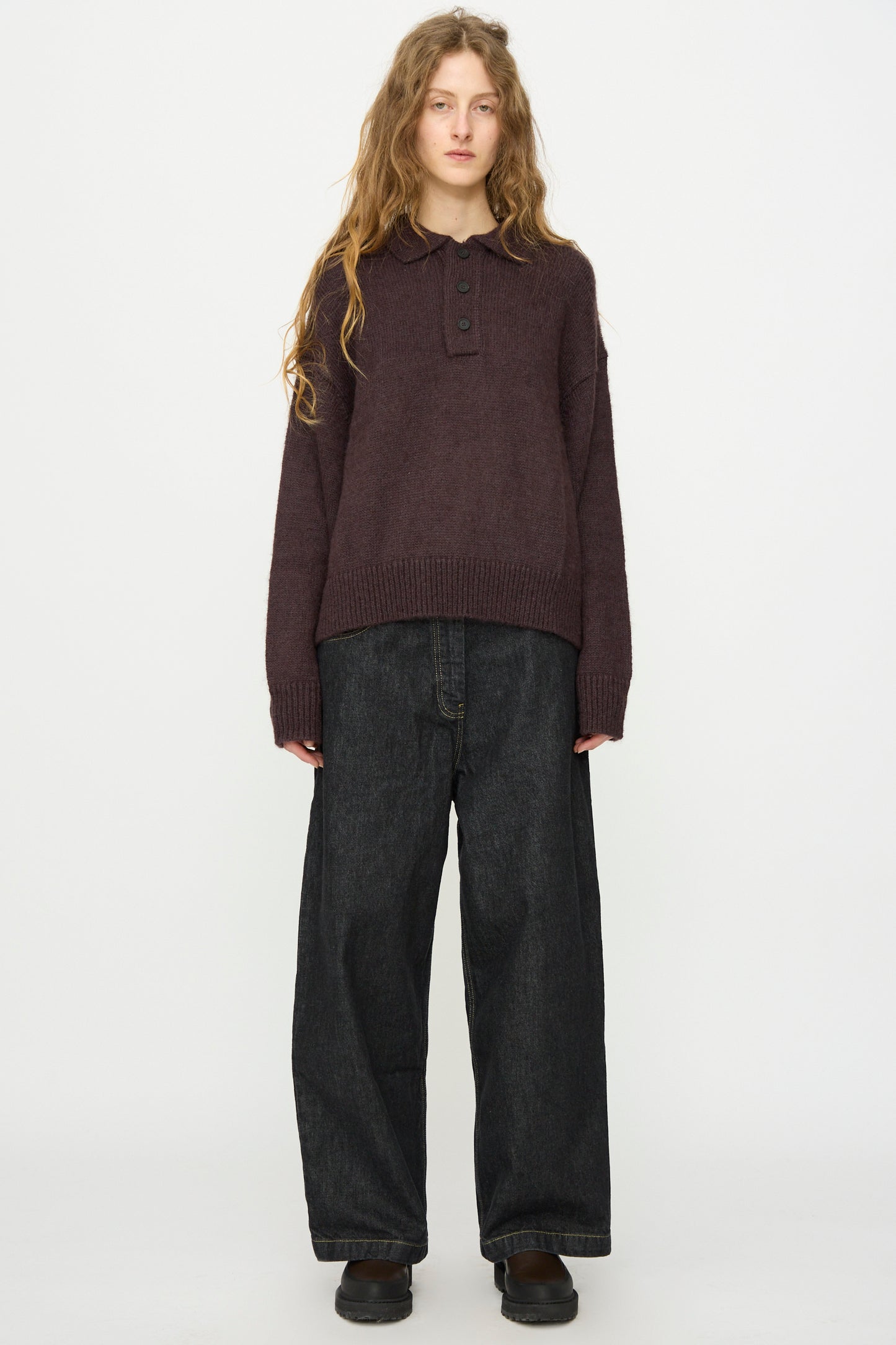 A person with long wavy hair wears a dark brown knit sweater with buttons, Studio Nicholson's Italian Denim Neive Pant in Black, and black shoes, standing against a plain white background.